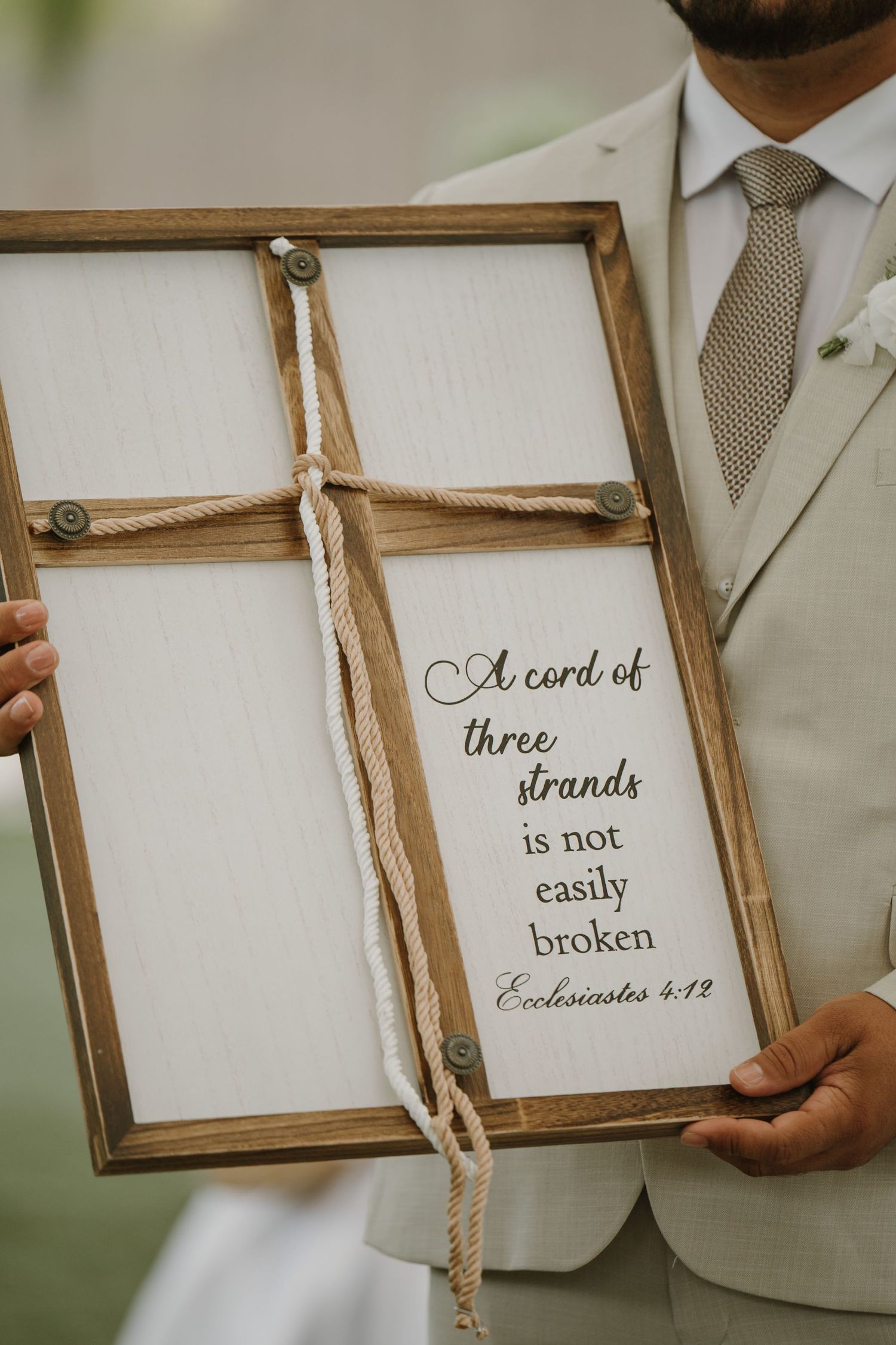 Groom holding a wooden framed sign with rope cross reading Ecclesiastes 4:12 cord of three strands wedding ceremony.