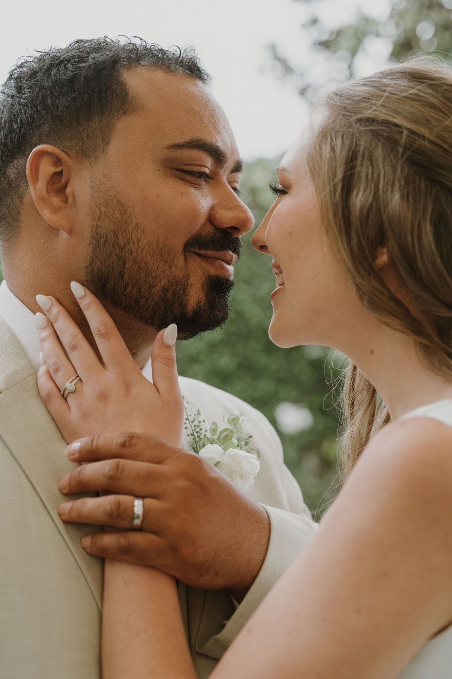 Smiling newlyweds share a tender moment, bride holding grooms face with wedding rings visible.