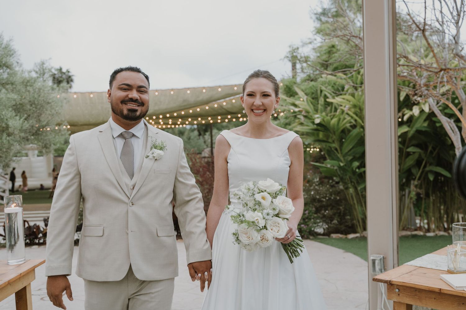 Smiling bride and groom holding hands at outdoor wedding ceremony, bride in white gown with bouquet, groom in beige suit.