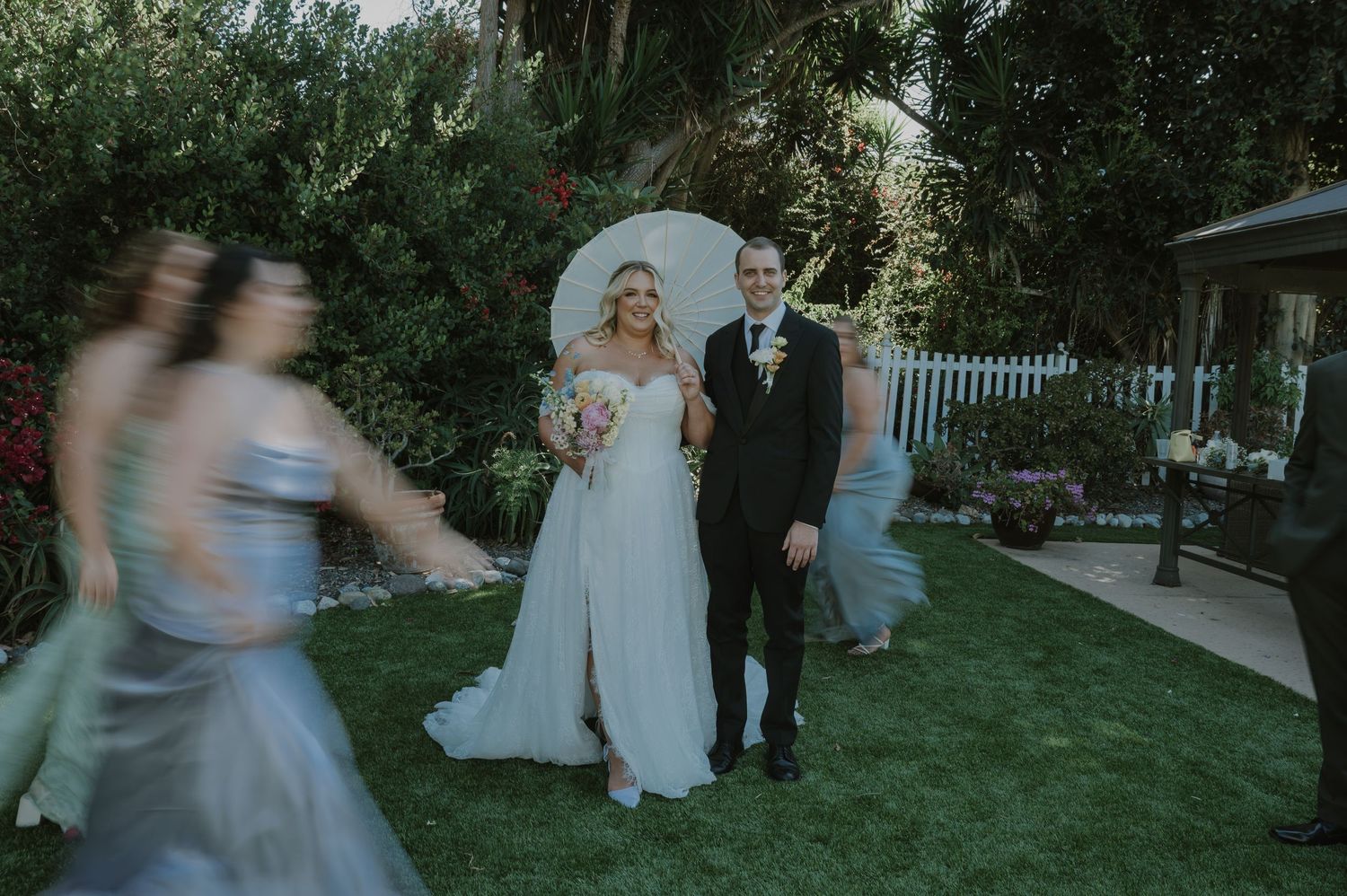 Bride and groom walk through a lush garden as bridesmaids pass by with white parasols.