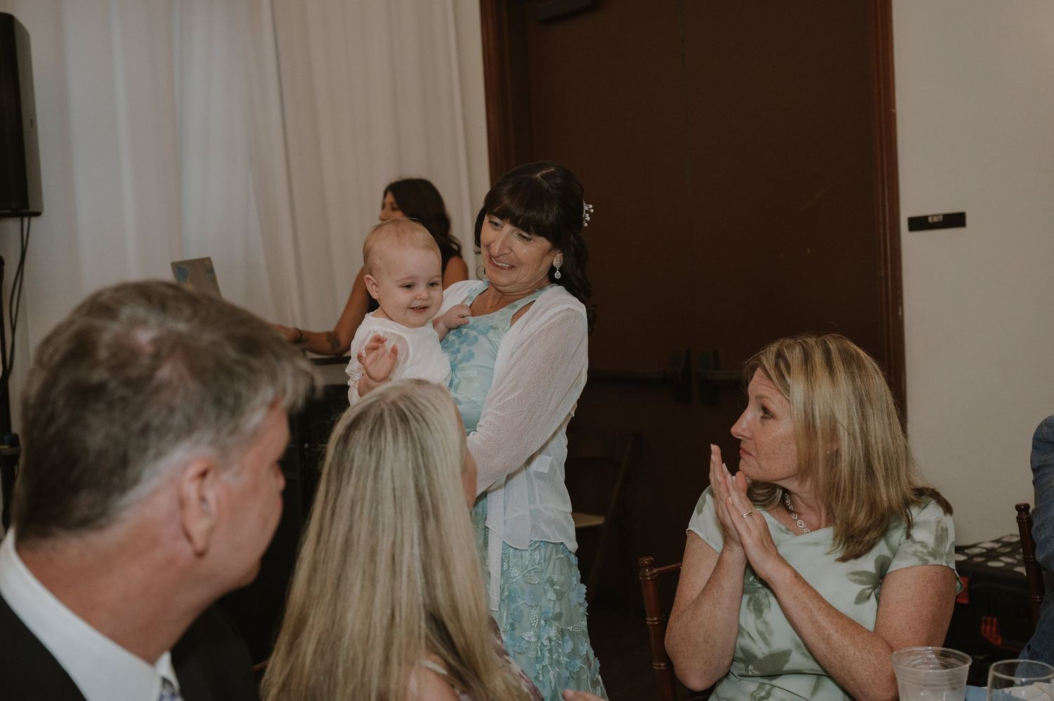 Woman holding a baby at a wedding reception while guests smile and clap in an elegant venue.