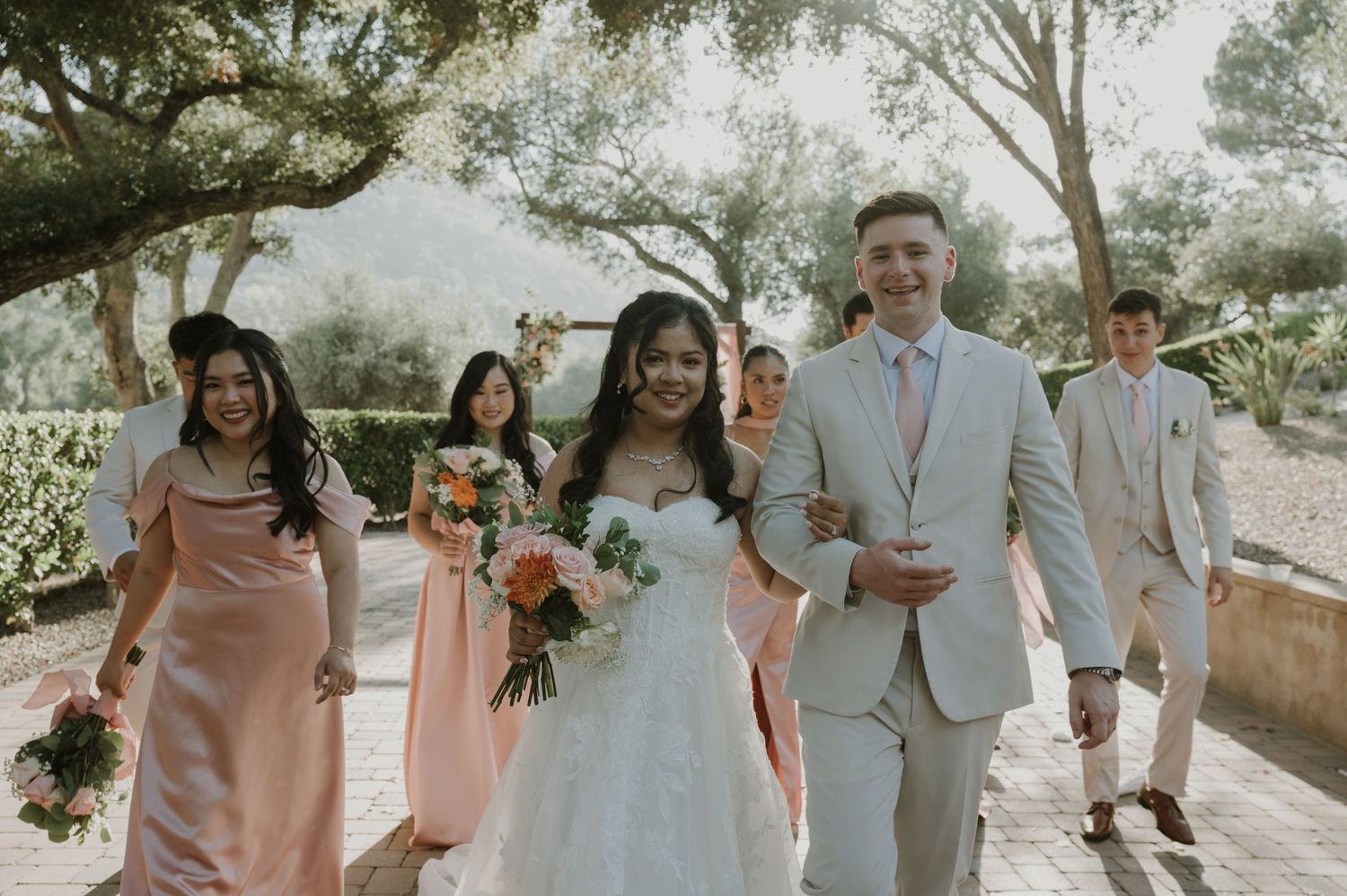 Bride in white gown walks with groom and bridesmaids in pink dresses at outdoor wedding.