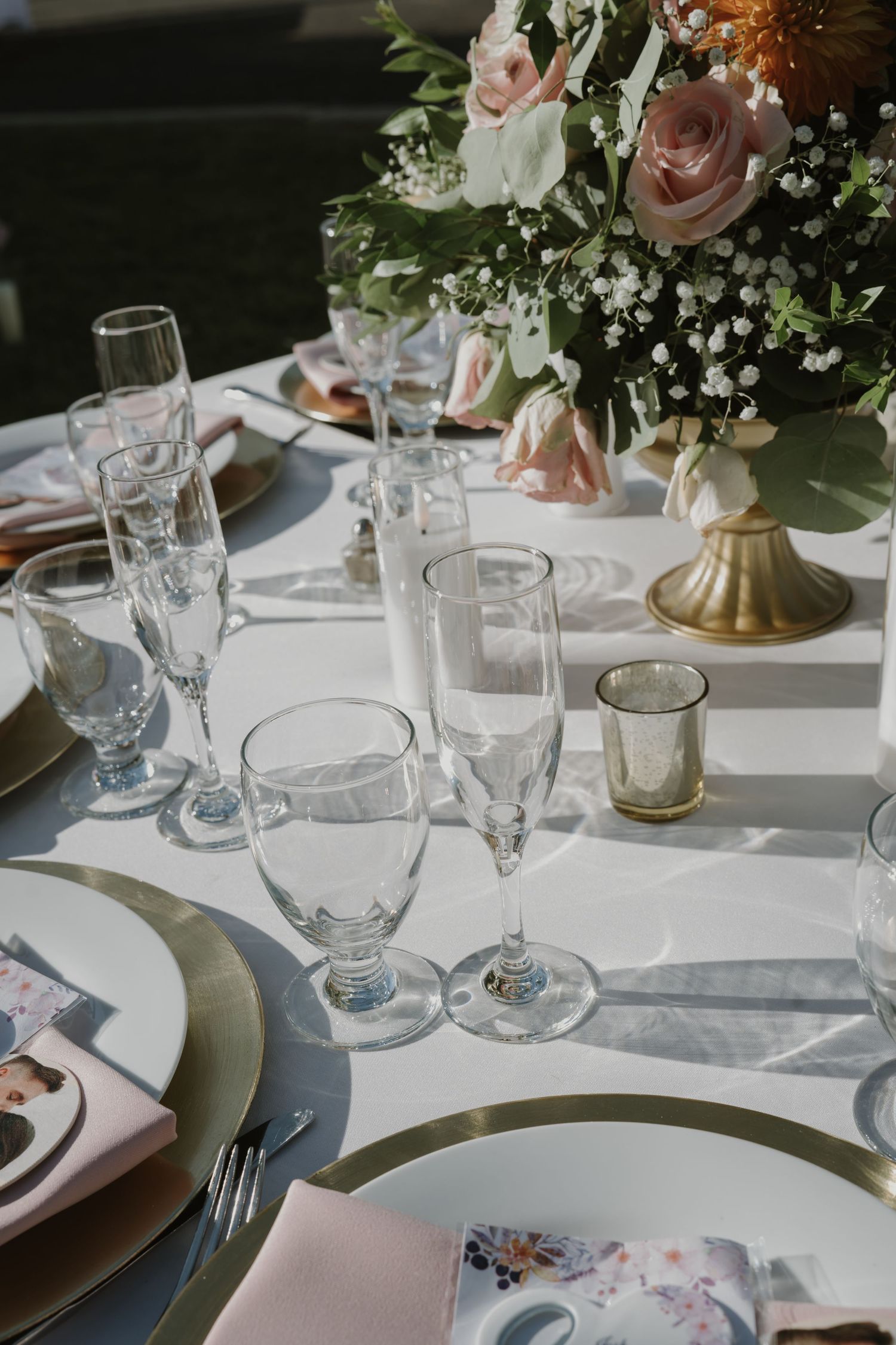 Elegant wedding reception table with champagne flutes, white plates, gold chargers, pink napkins, and a floral centerpiece.