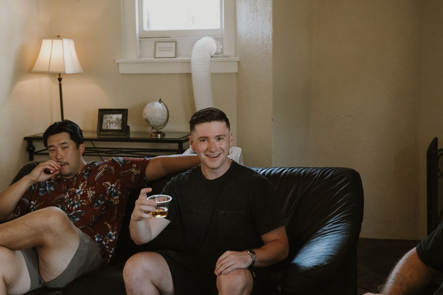 Two young men relaxing on a leather couch in a cozy living room, one holding a small cup and smiling.
