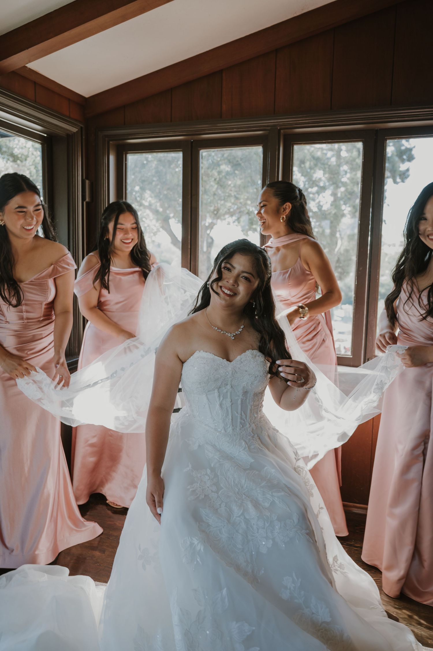 Bride in white lace gown smiles as bridesmaids in pink dresses hold her veil in a wood-paneled room.