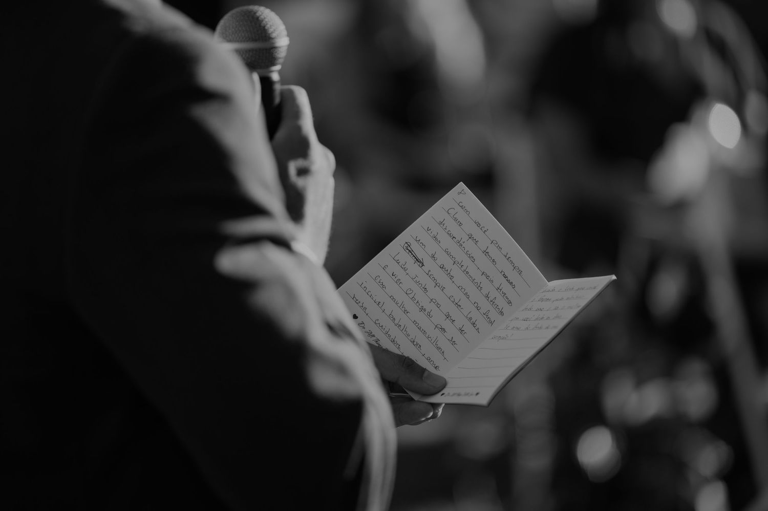 A person reads wedding vows from a booklet while holding a microphone at a ceremony in black and white.