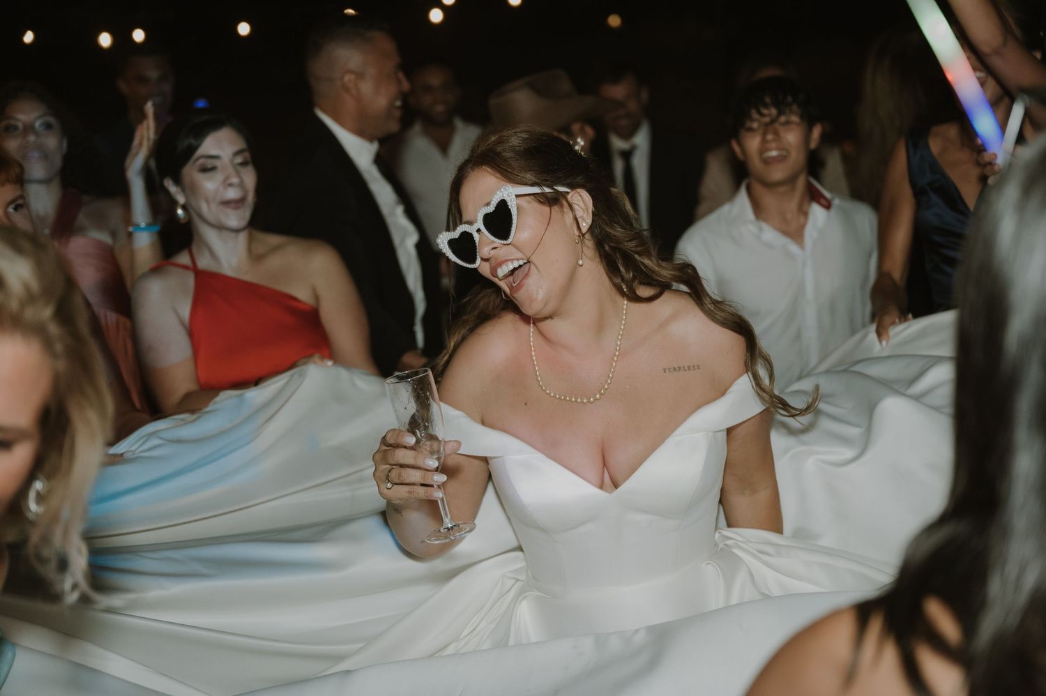 Bride in white off-shoulder gown and heart sunglasses dancing joyfully at wedding reception.