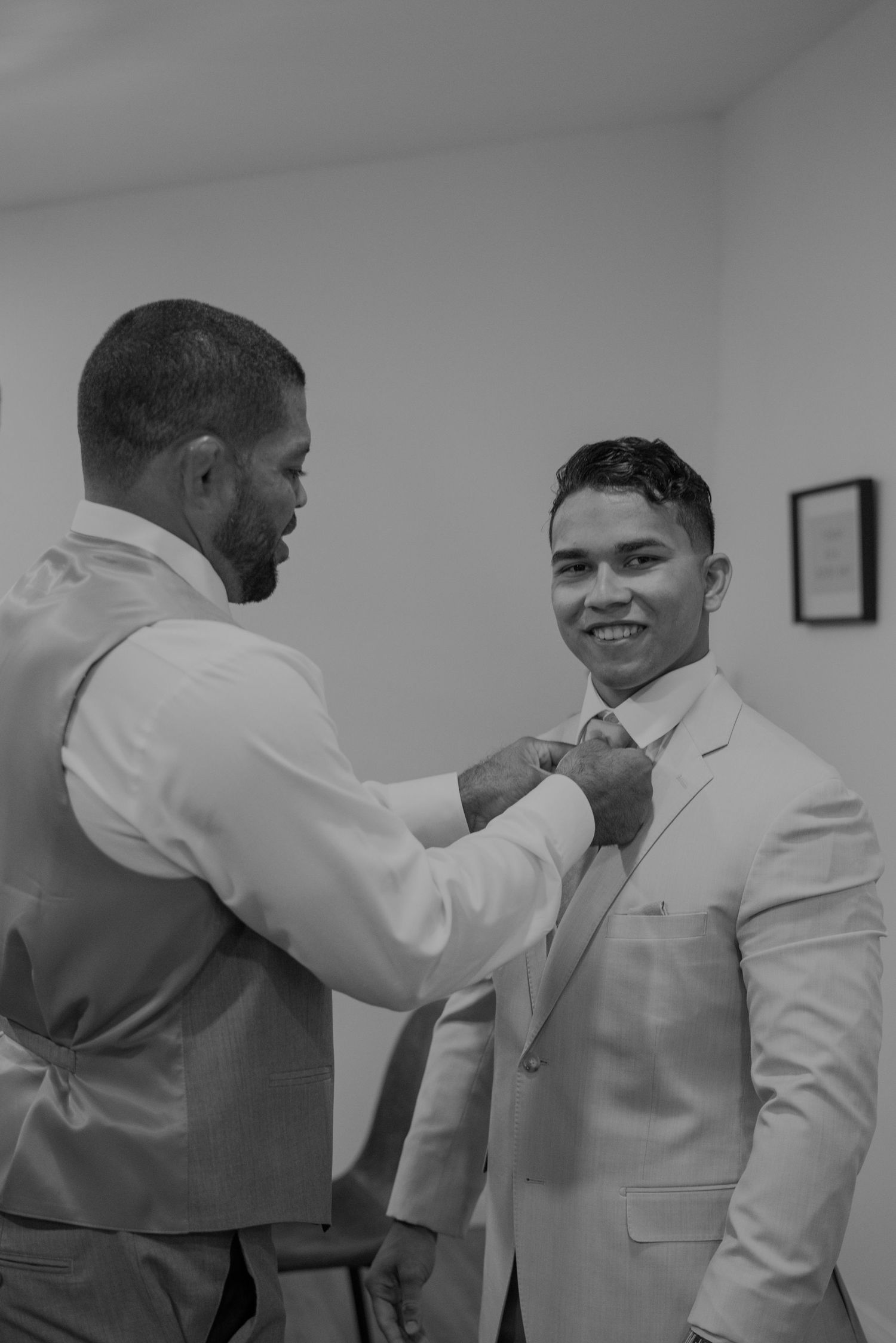 Best man helping smiling groom straighten his tie in black and white wedding preparation photo.