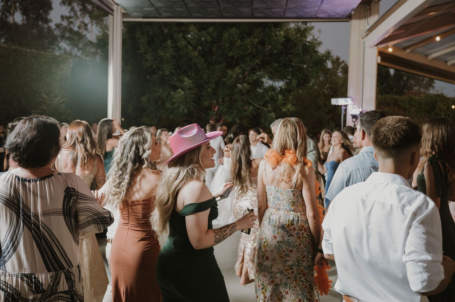 Wedding guests dancing outdoors at night, woman in pink cowboy hat stands out in the crowd.