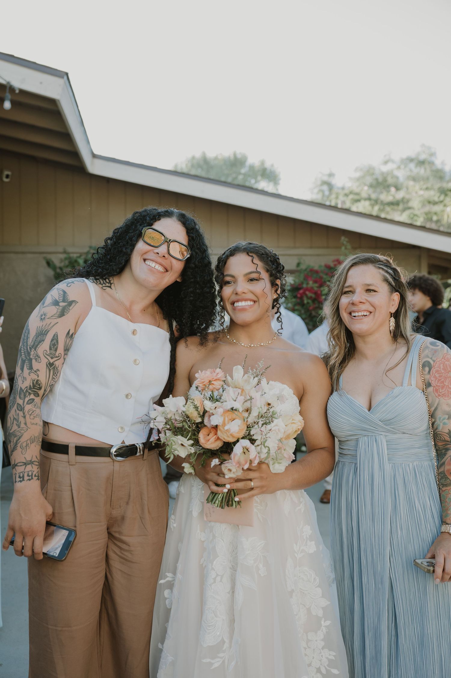 Bride holding peach bouquet posing with two smiling guests at outdoor wedding.