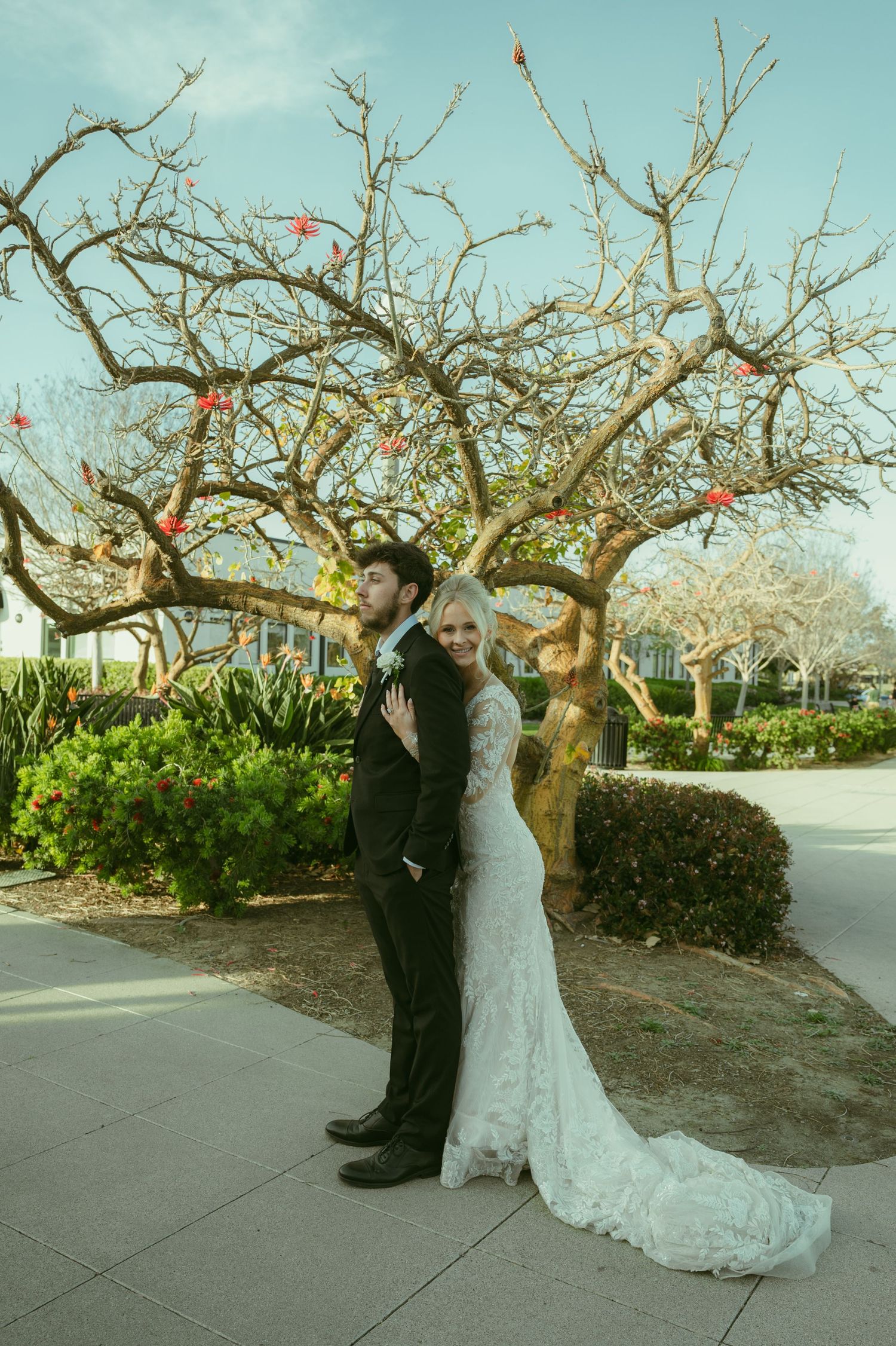 Bride in lace gown and groom in black suit pose back-to-back before a bare tree with red blooms outdoors.