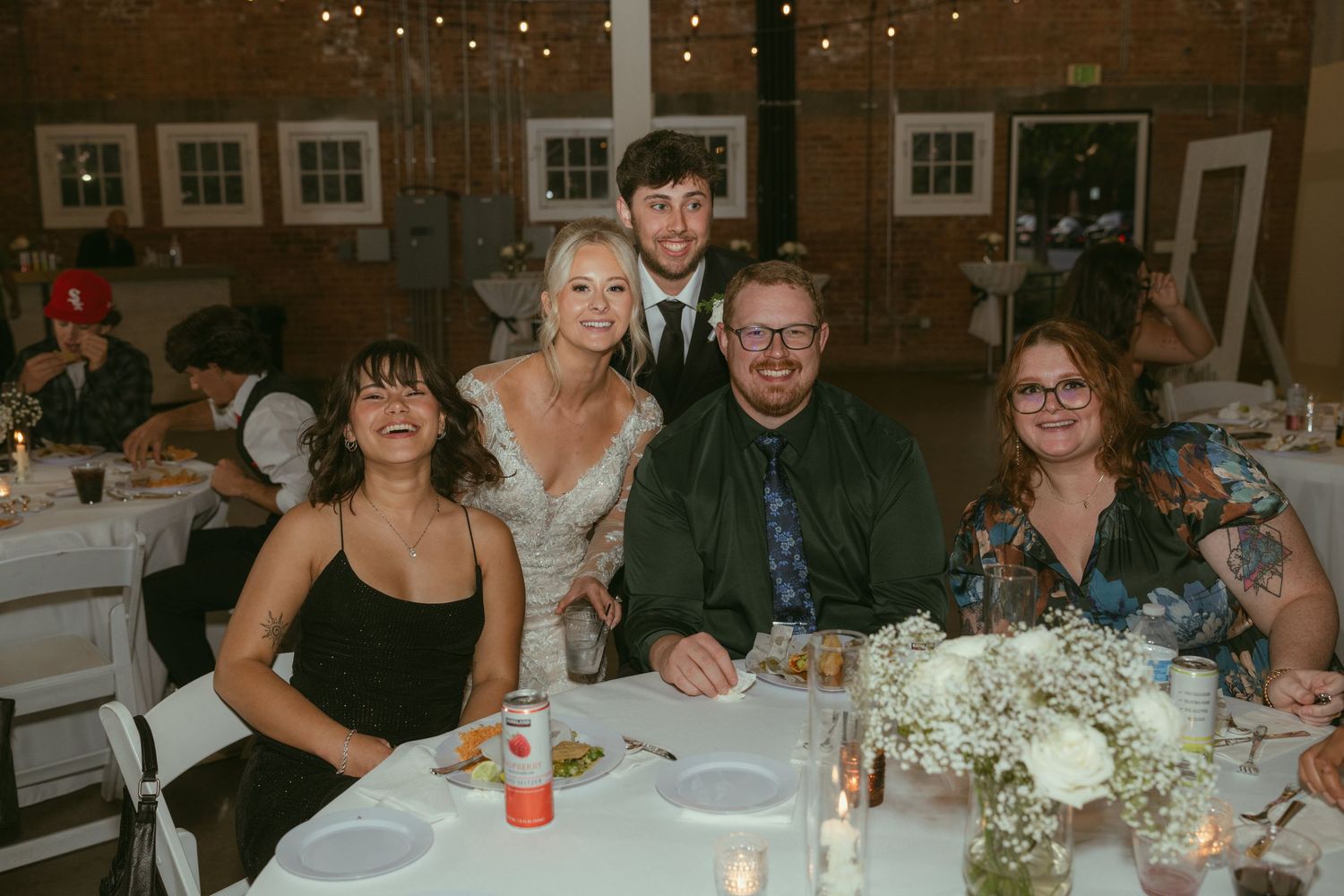 Wedding guests smile at a reception table decorated with candles and white floral centerpieces indoors.