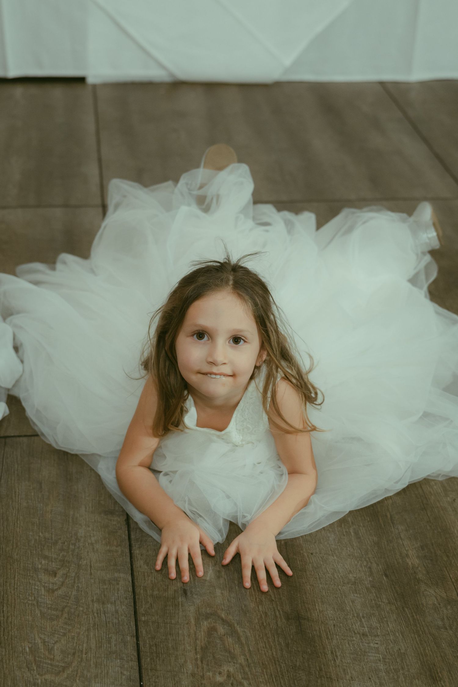 Adorable young flower girl in a white tulle dress sitting on a wooden floor looking up.