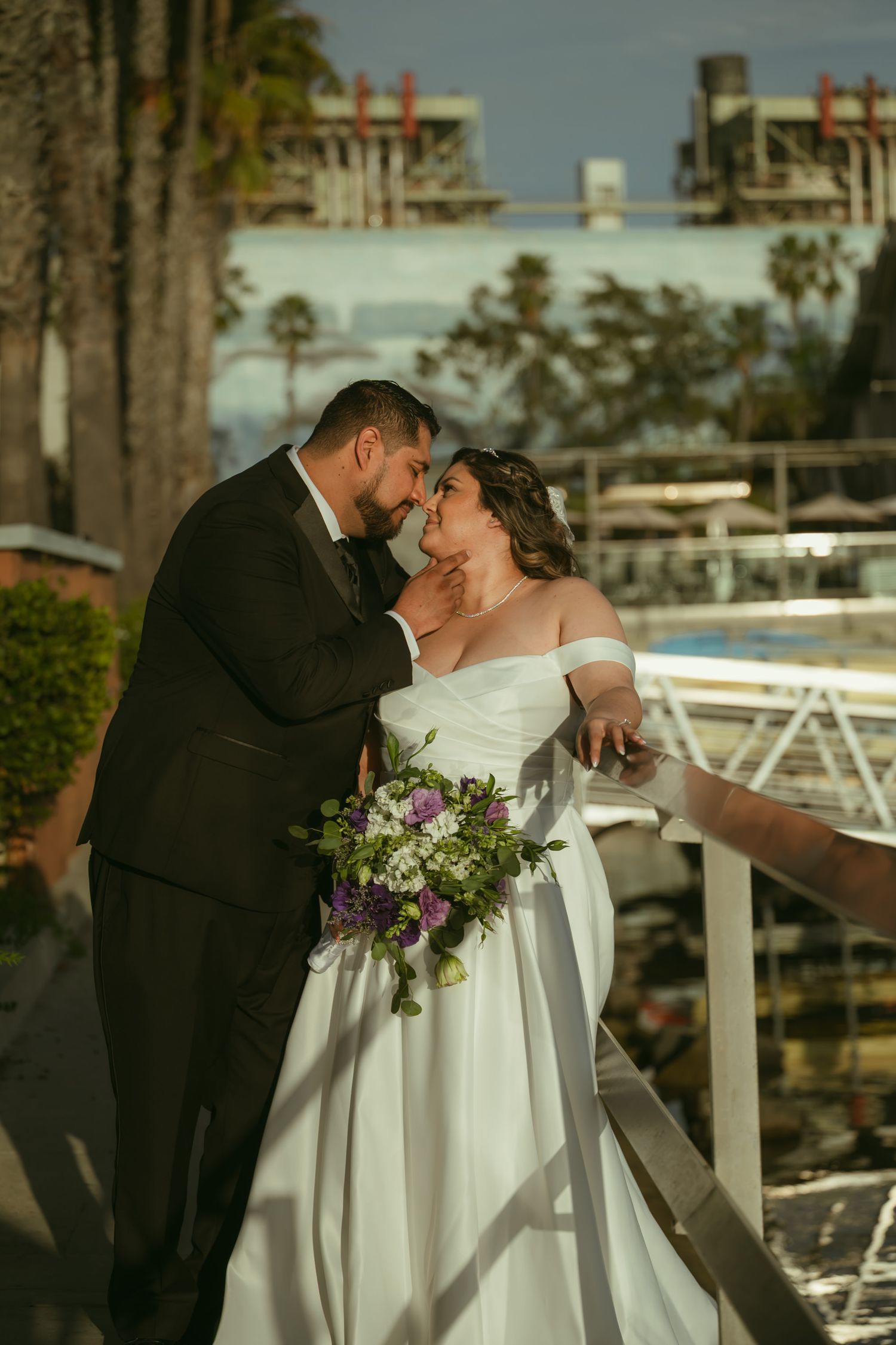 Bride and groom in wedding attire share a romantic moment on a balcony with palm trees behind.