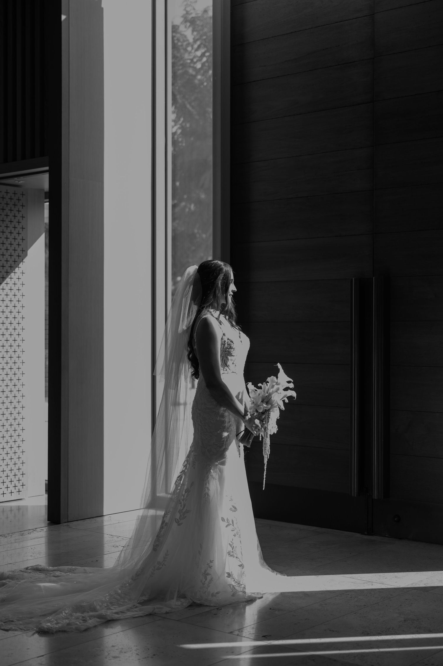 Bride in elegant lace wedding gown and veil holds bouquet, standing by sunlit window in black and white portrait.
