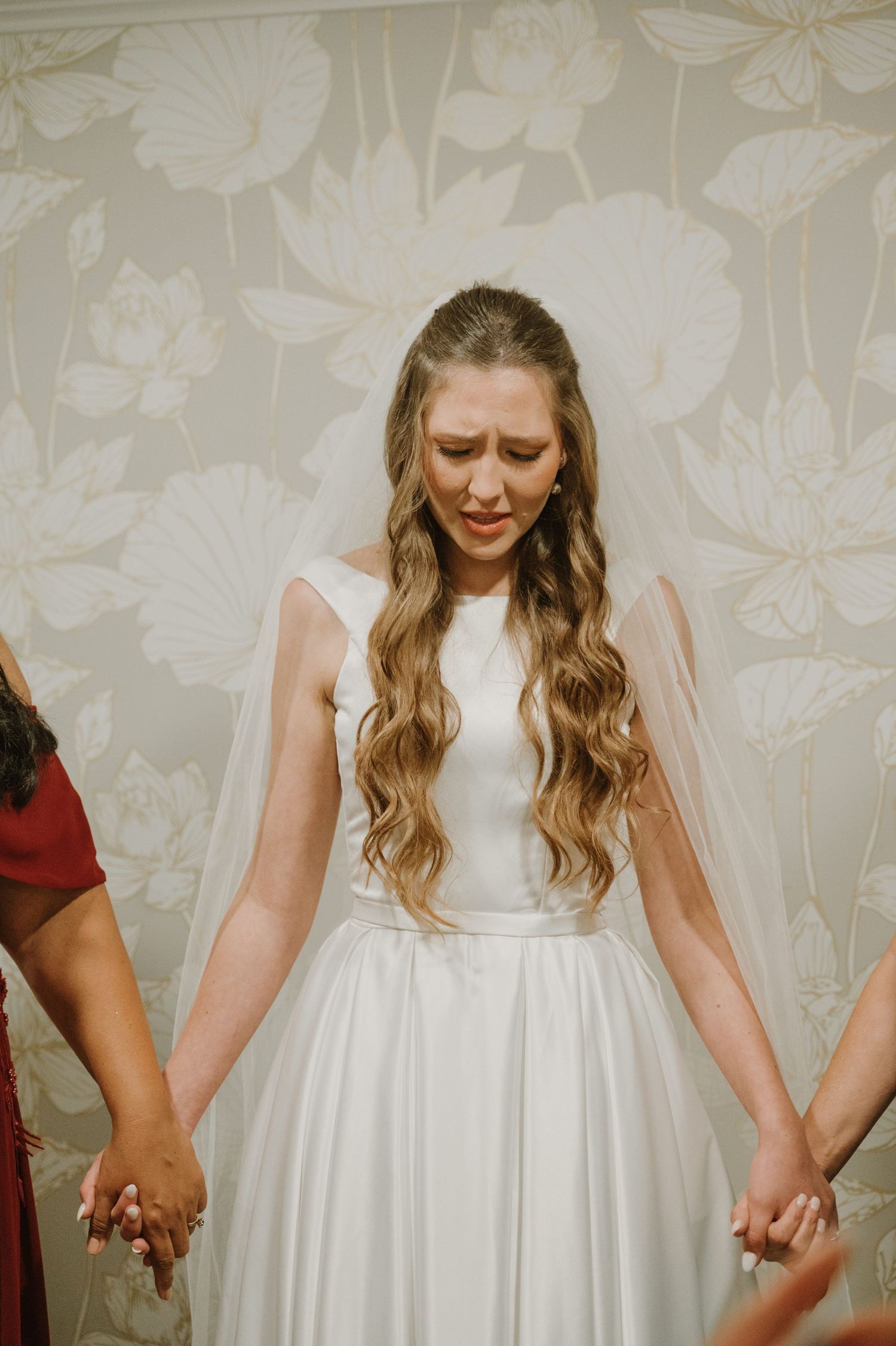 Bride in white gown holding hands with bridesmaids, eyes closed in prayer, standing before floral wallpaper.