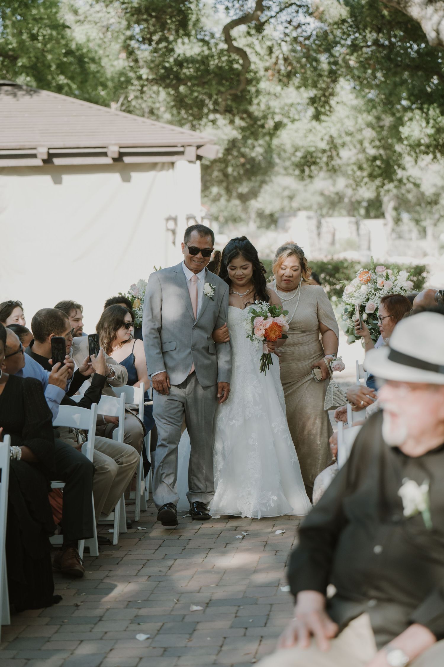 Bride walks down the aisle escorted by two family members at an outdoor wedding ceremony.