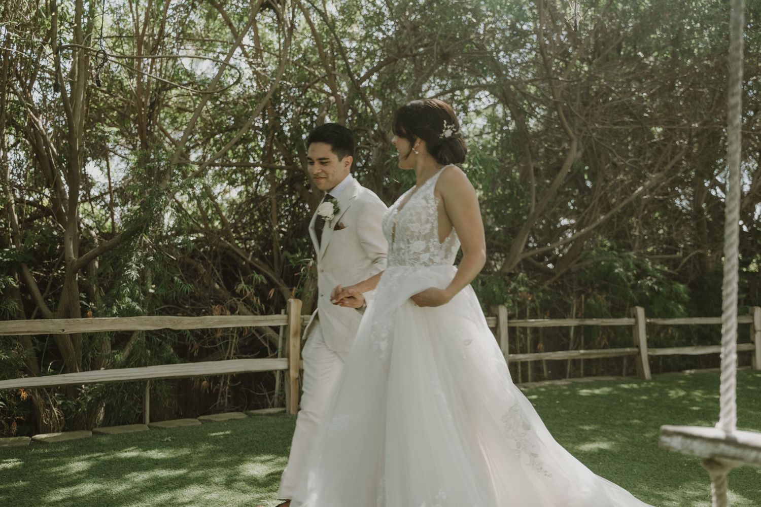A couple in white wedding attire walks together outdoors, surrounded by lush green trees and a wooden fence.