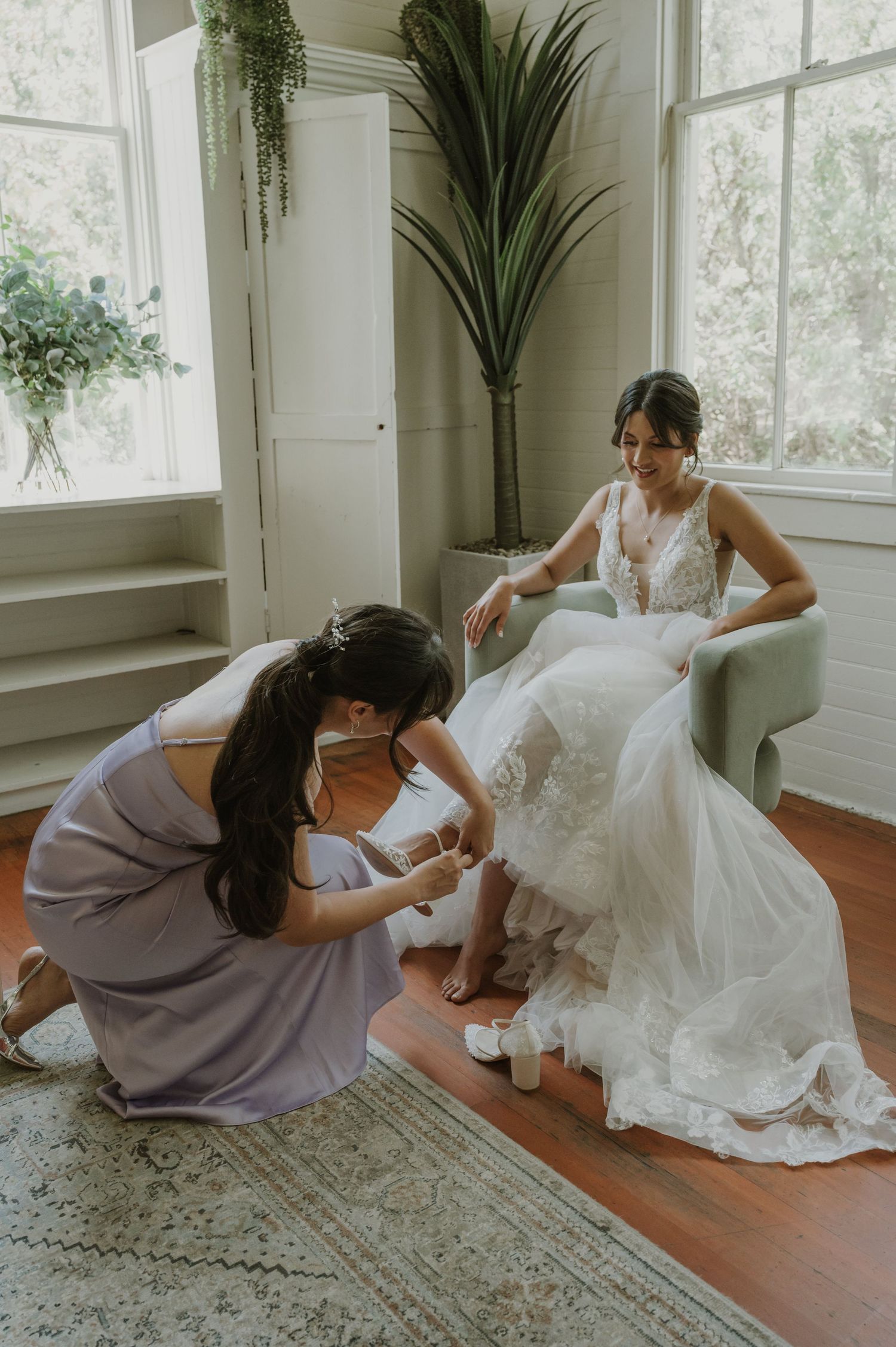 Bridesmaid in lavender dress helping bride put on wedding shoes in bright white room.