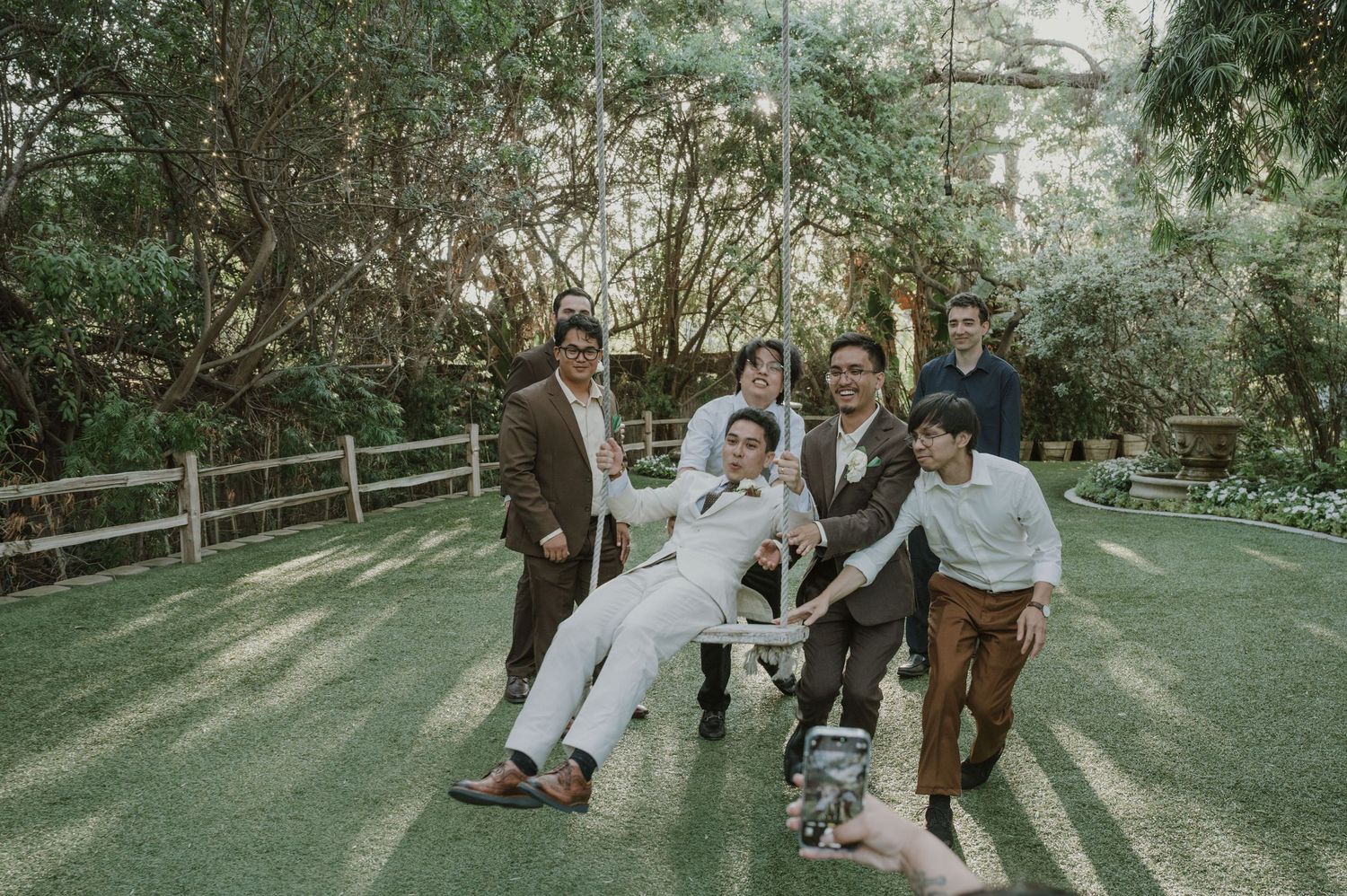 Groom seated in chair lifted by groomsmen in suits outdoors at garden wedding venue.