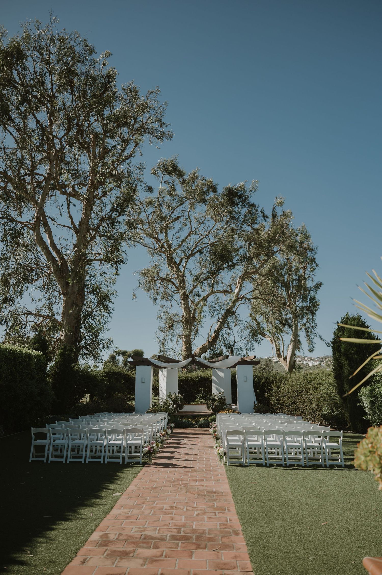 Outdoor wedding ceremony setup with white chairs, brick aisle, floral arch, and tall trees under a clear blue sky.