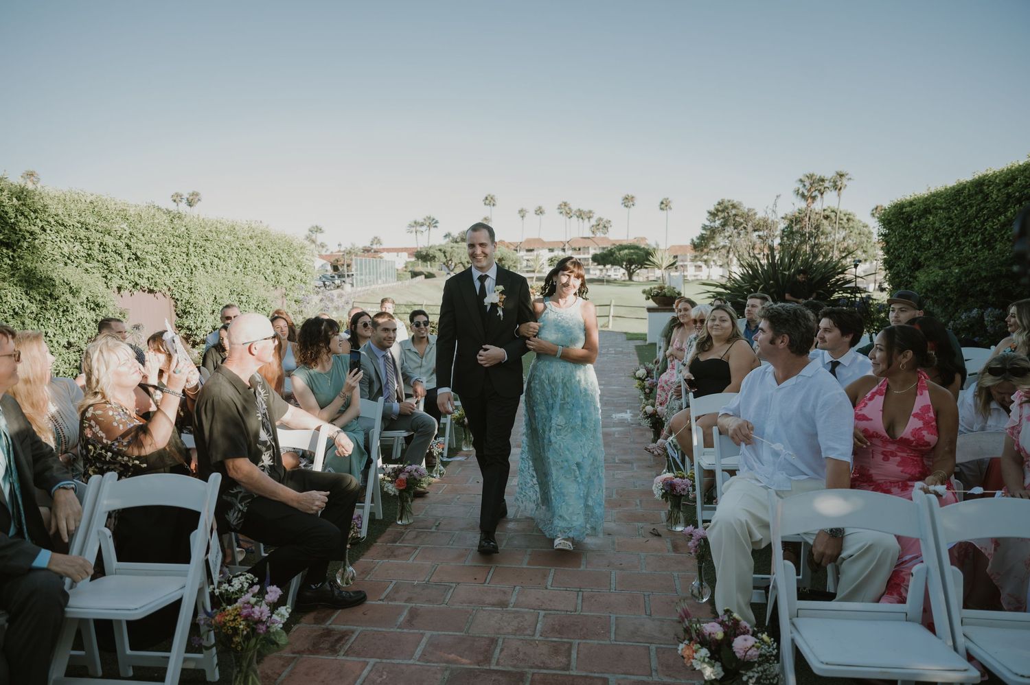 Bridesmaid in blue gown walks down outdoor wedding aisle with groomsman, guests seated on white chairs.