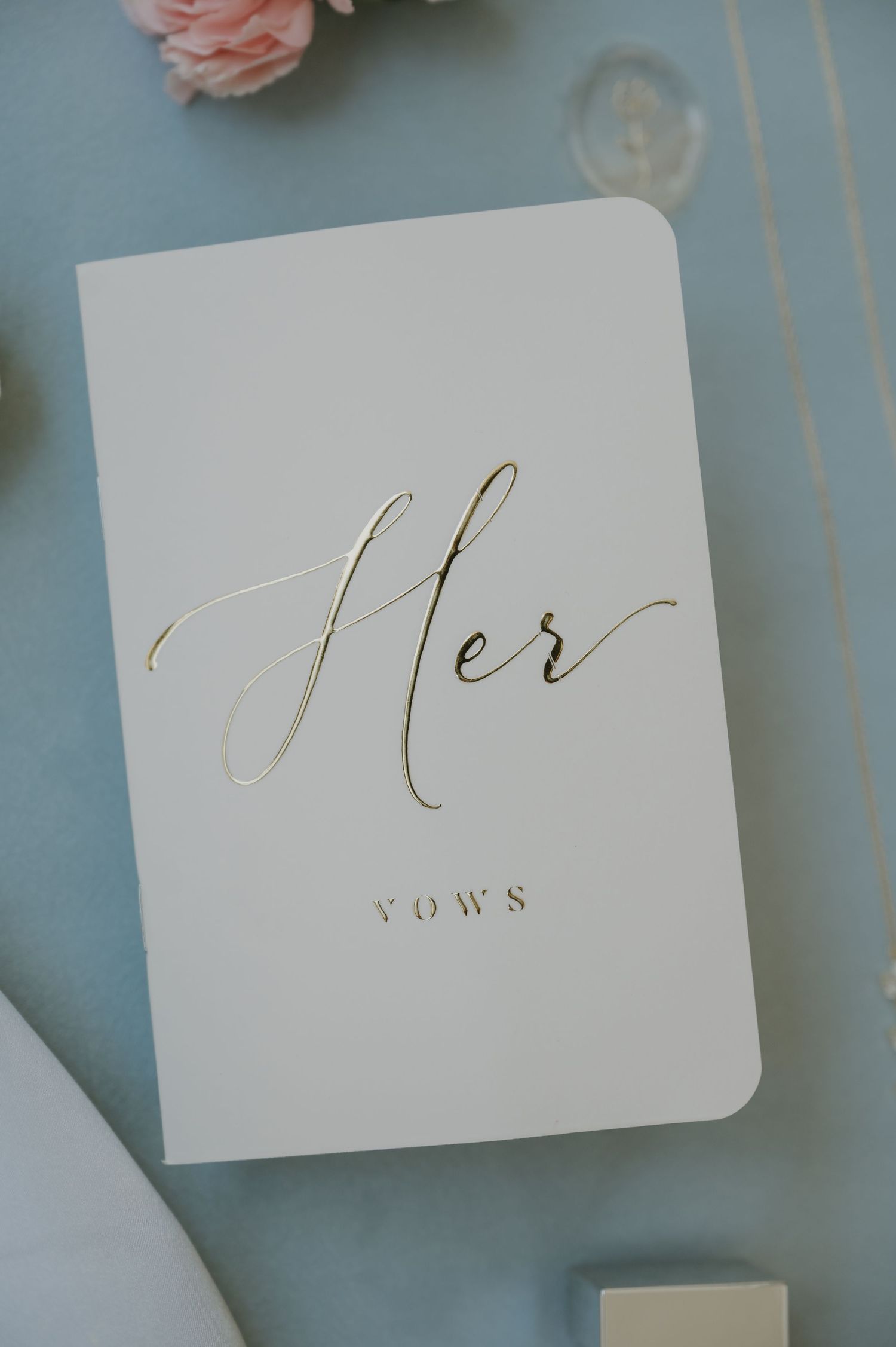 White wedding vow book with gold foil script reading Her Vows on a soft blue background with pink flowers.