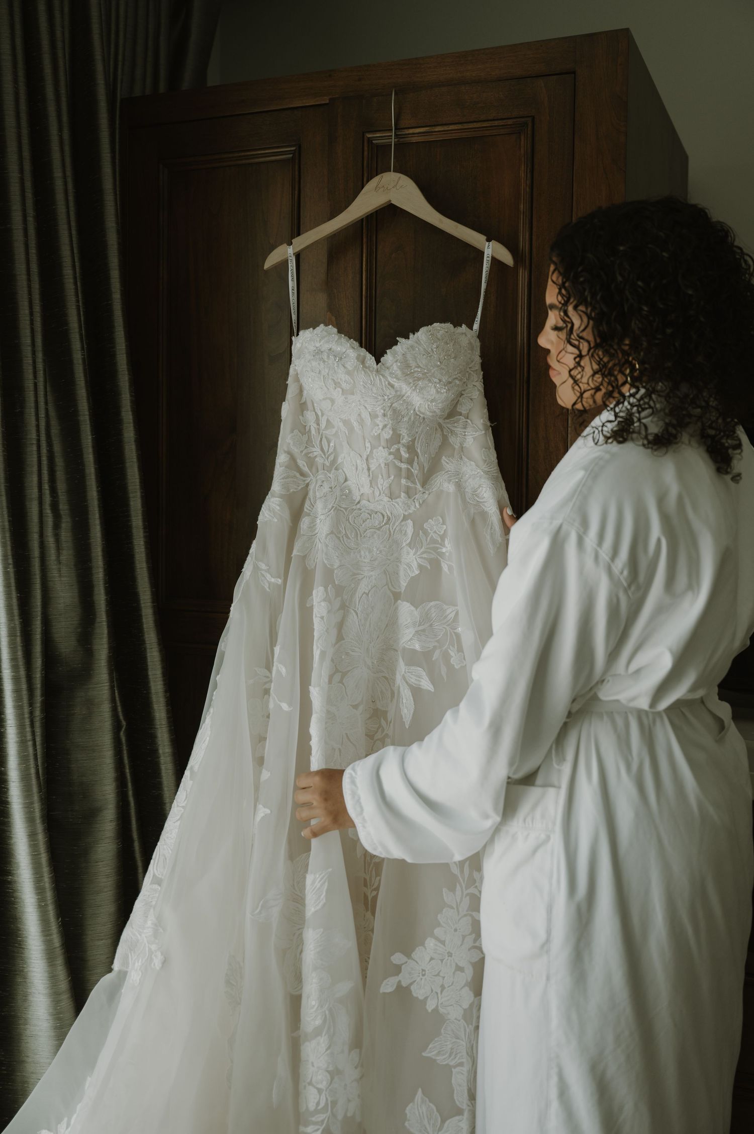 Bride in white robe admiring her lace strapless wedding gown hanging on a wooden wardrobe door.