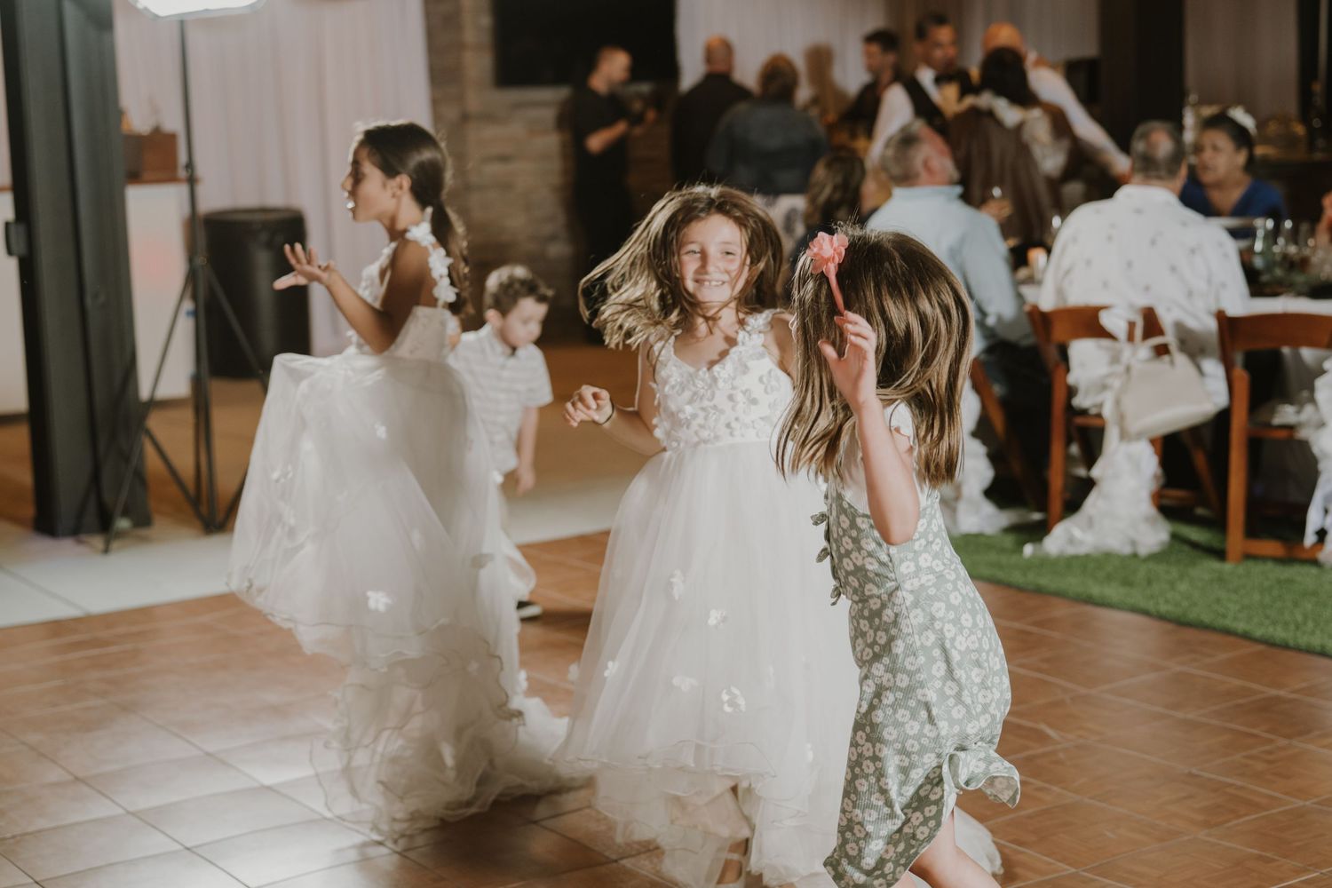 Young flower girls in white dresses dancing joyfully at a wedding reception on a tile dance floor.