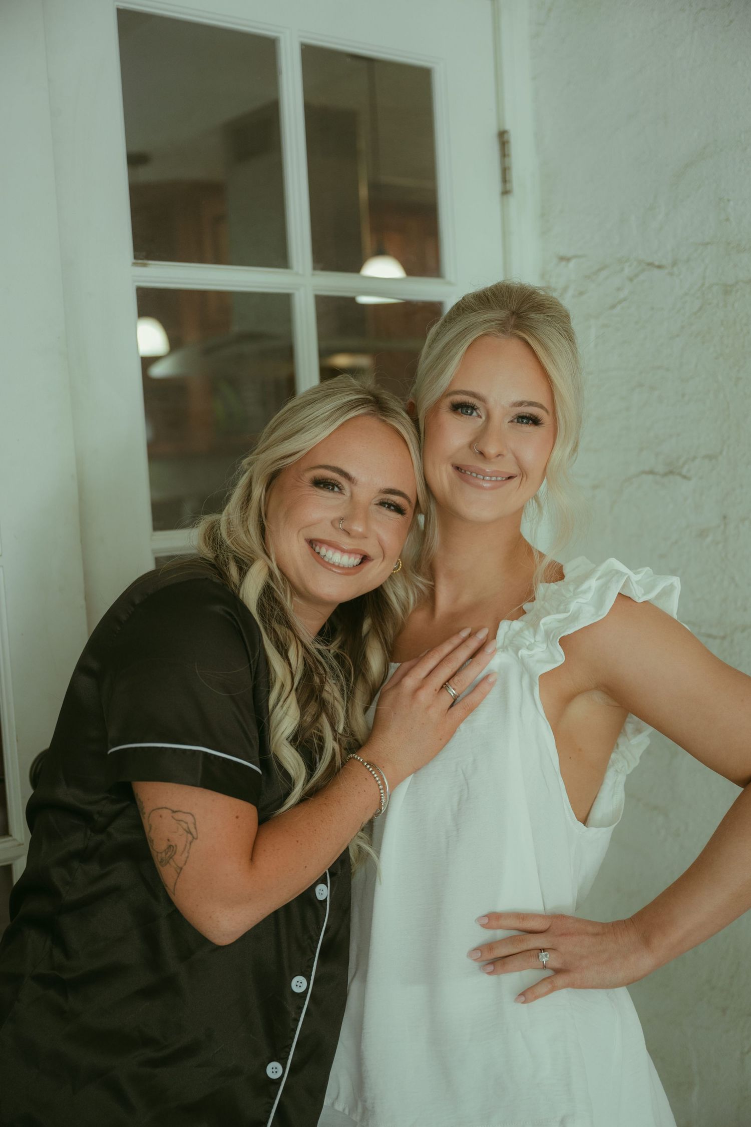 Two smiling blonde women pose together, one in black satin pajamas and one in a white ruffled dress, near a white brick wall.