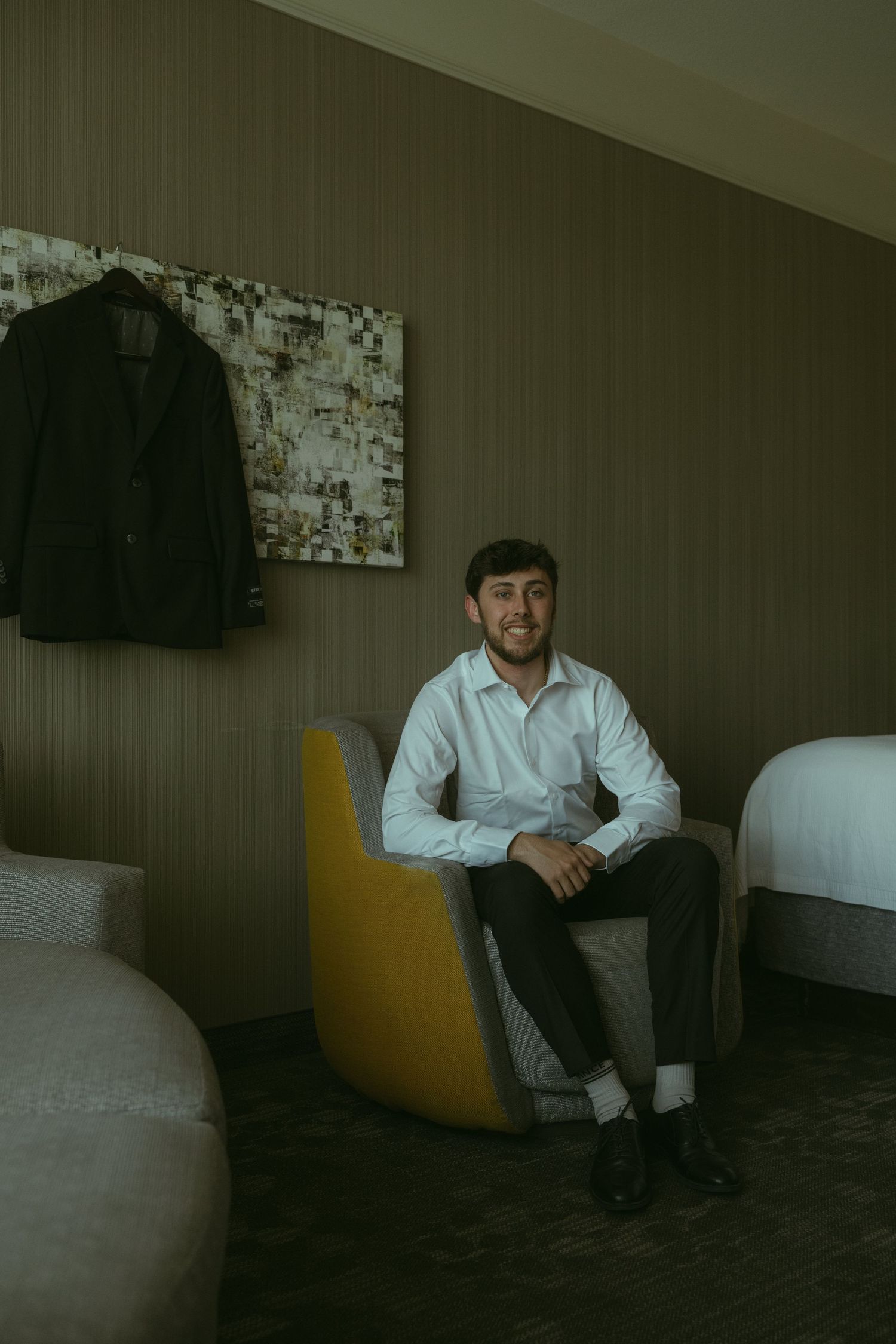 Groom sitting in yellow chair wearing white dress shirt and dark pants in a hotel room before wedding.