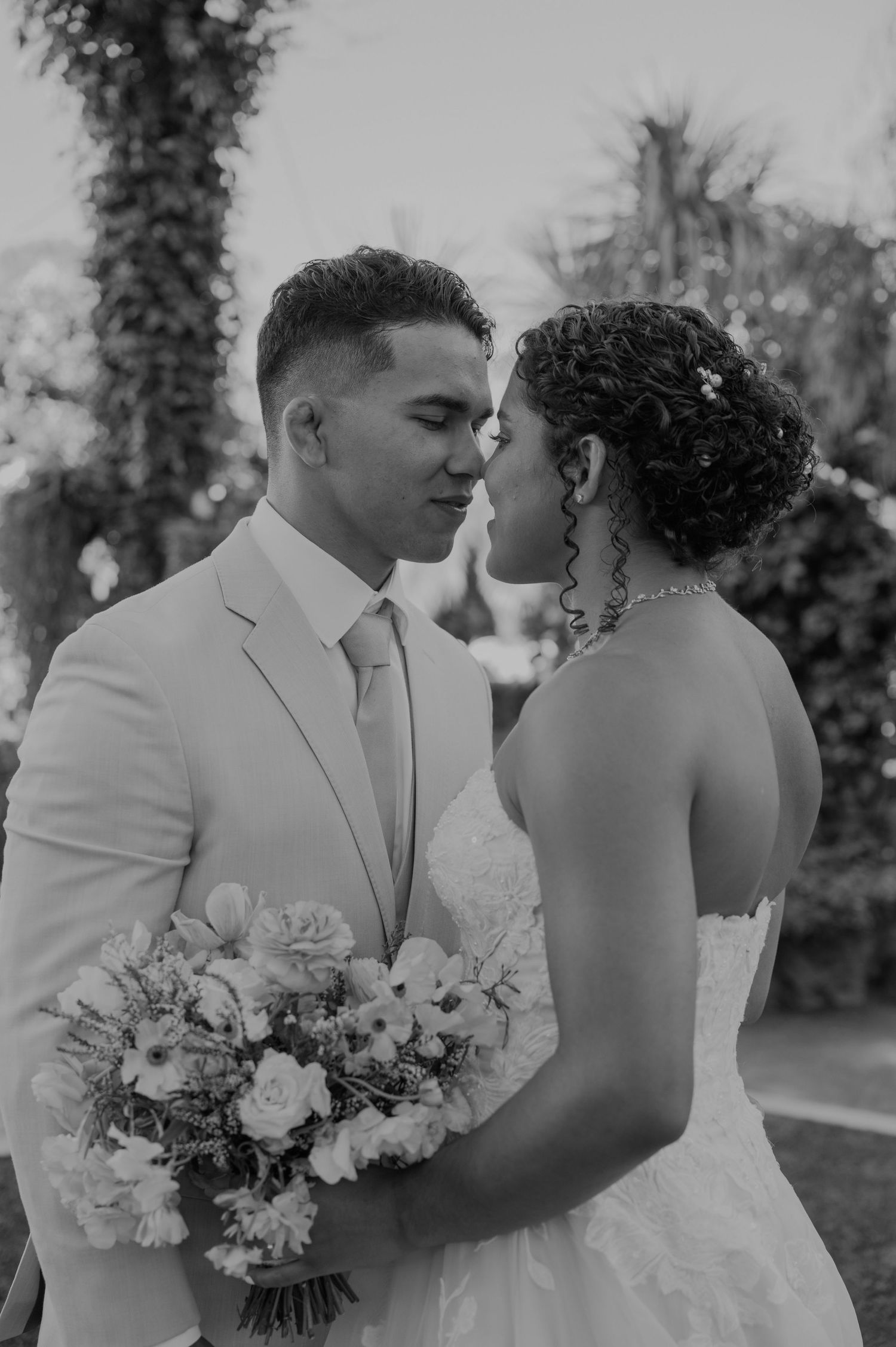 Black and white photo of a couple in wedding attire sharing an intimate moment, bride holding a floral bouquet outdoors.