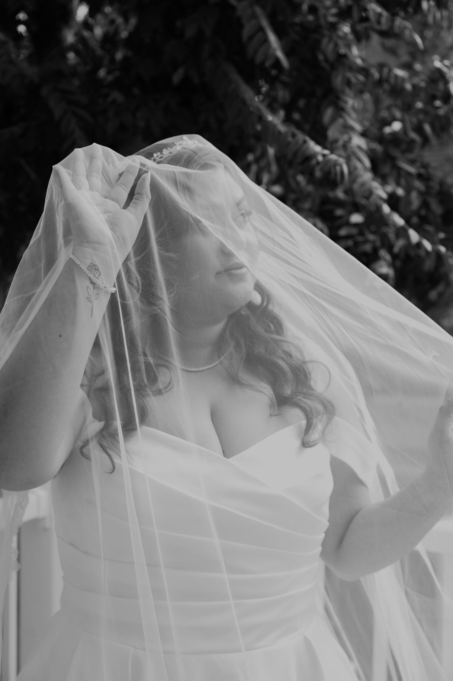 Black and white portrait of bride lifting sheer veil over face in strapless wedding dress.