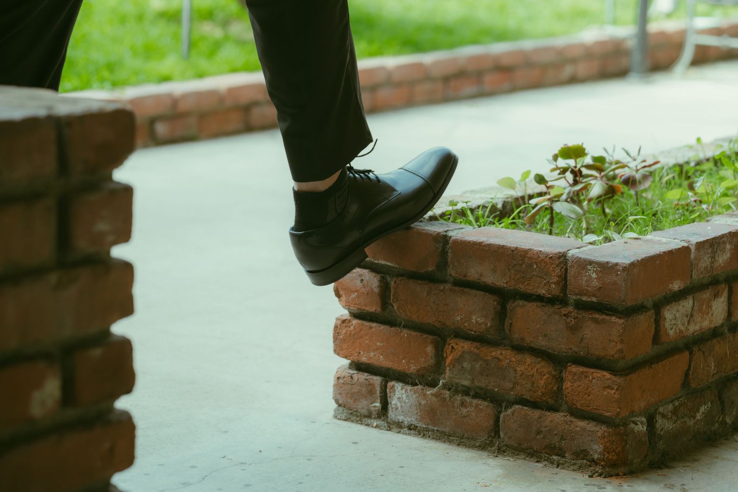 Person in black dress shoes and trousers stepping over a rustic red brick wall outdoors.