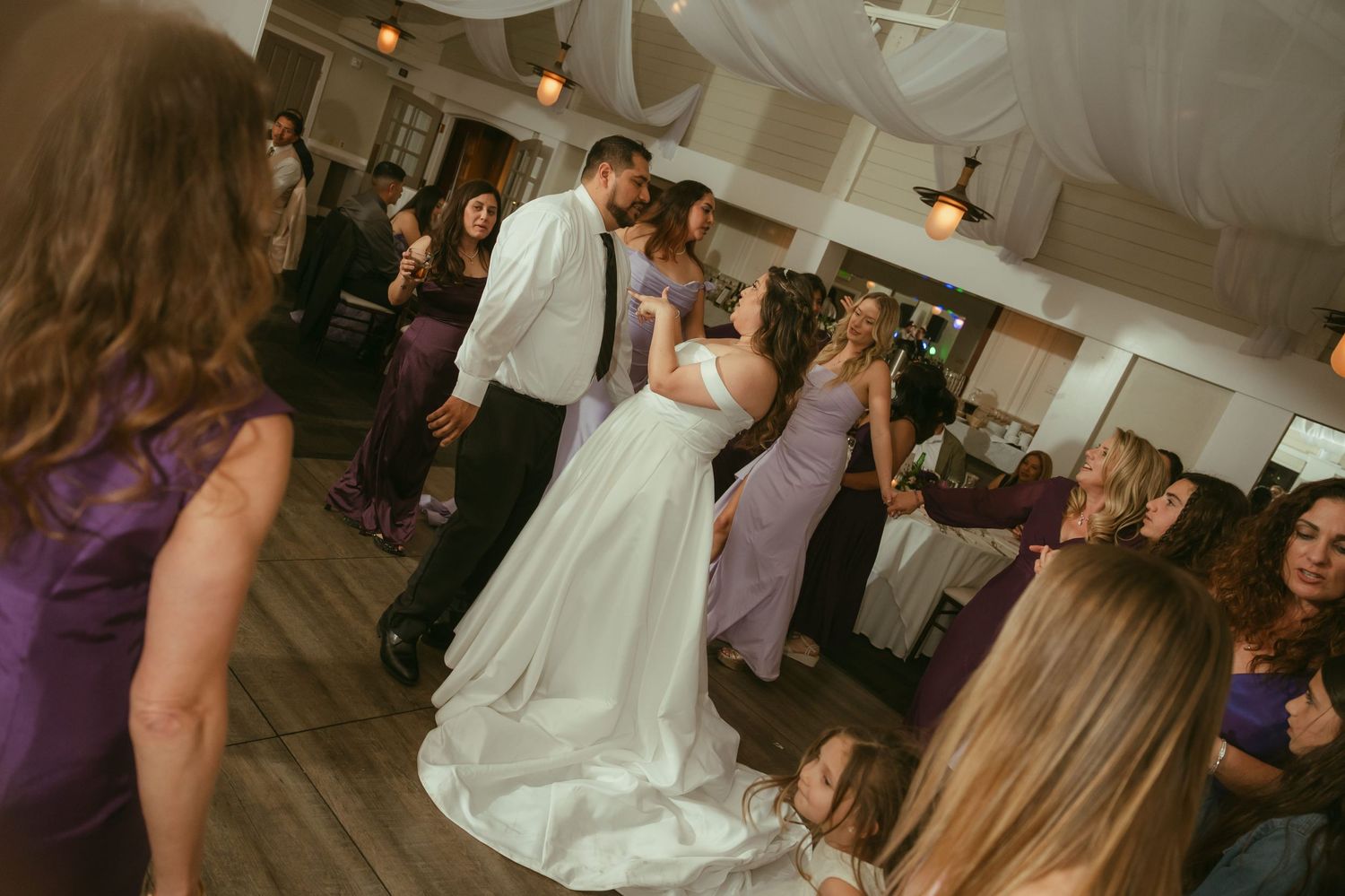 Bride and groom dance together surrounded by guests in purple dresses at lively wedding reception.