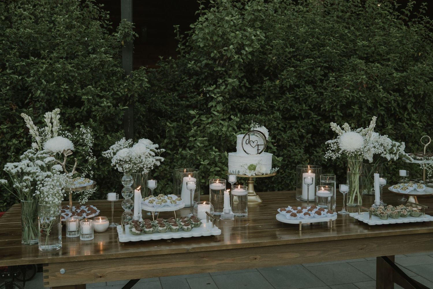 Elegant wedding dessert table with white floral arrangements, candles, and tiered treats on a rustic wood table outdoors.