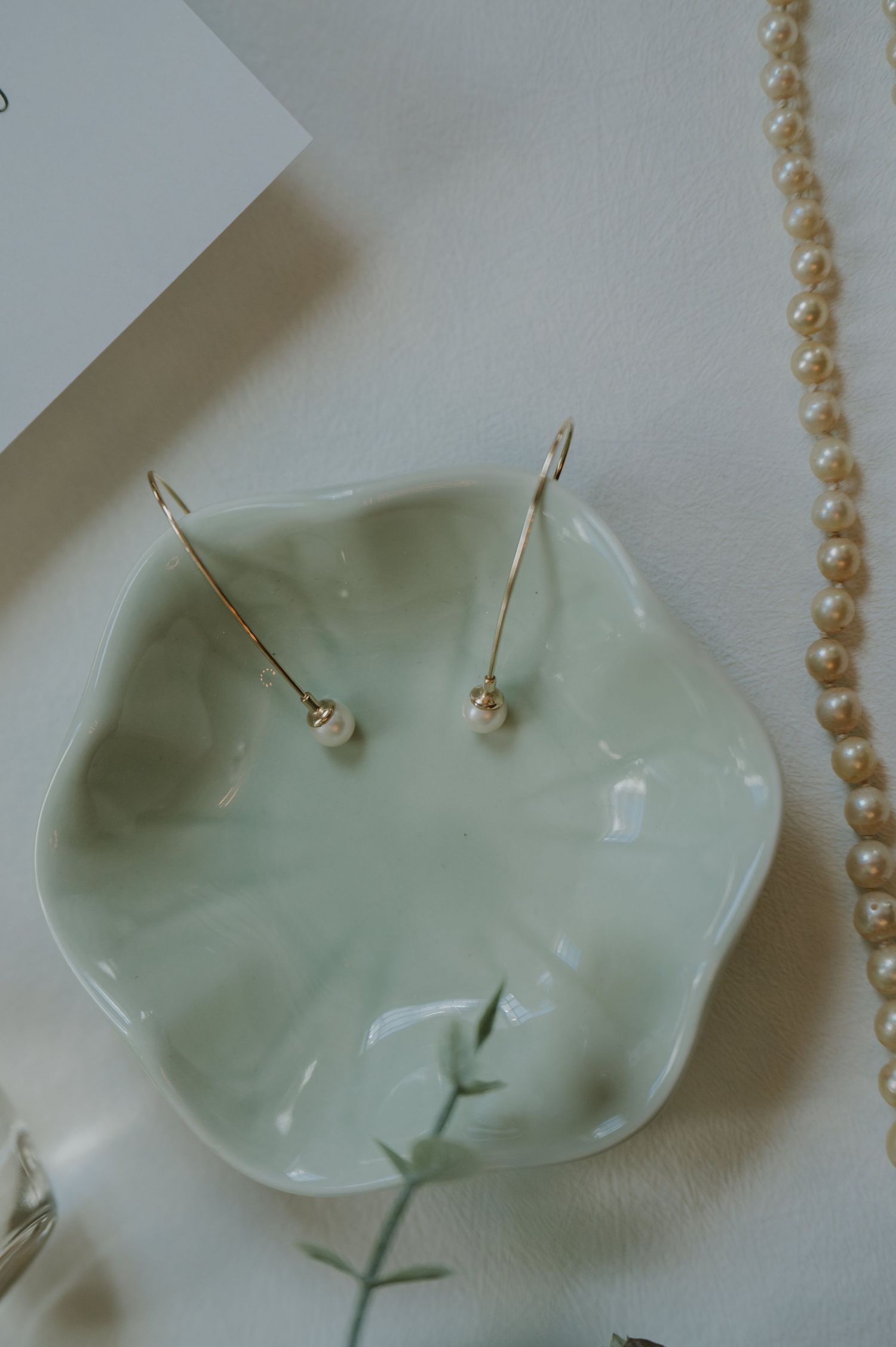 Gold drop pearl earrings resting in a mint green ceramic dish beside a pearl necklace and eucalyptus.