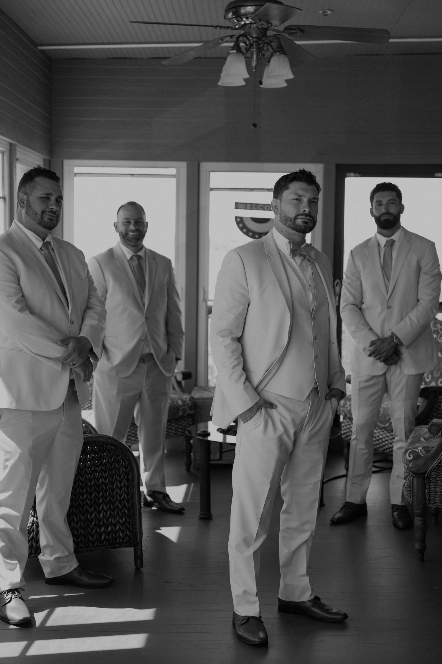 Group of groomsmen in light colored suits standing in sunlit room.