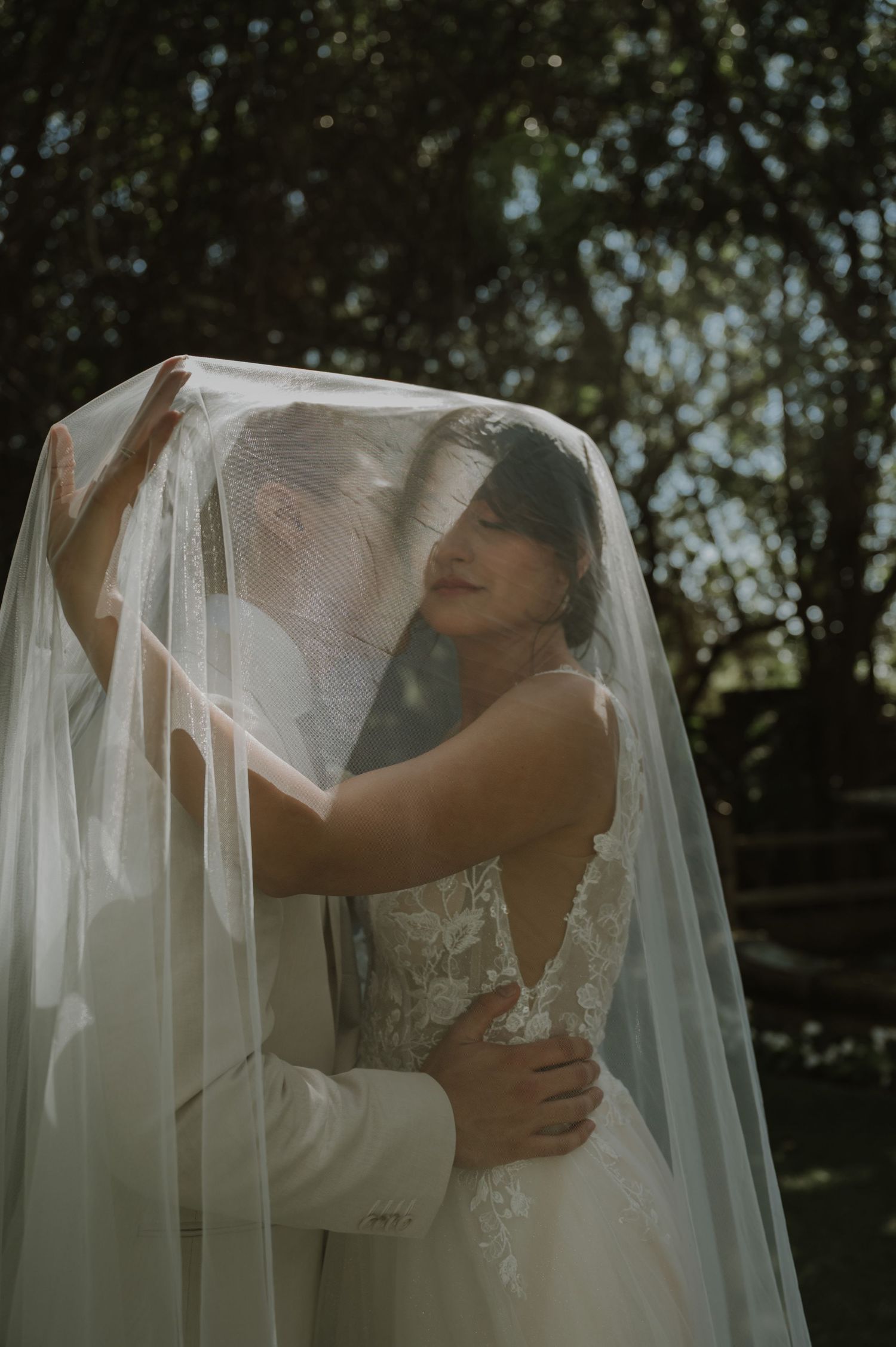 Romantic wedding portrait of couple embracing under flowing white veil in garden setting.