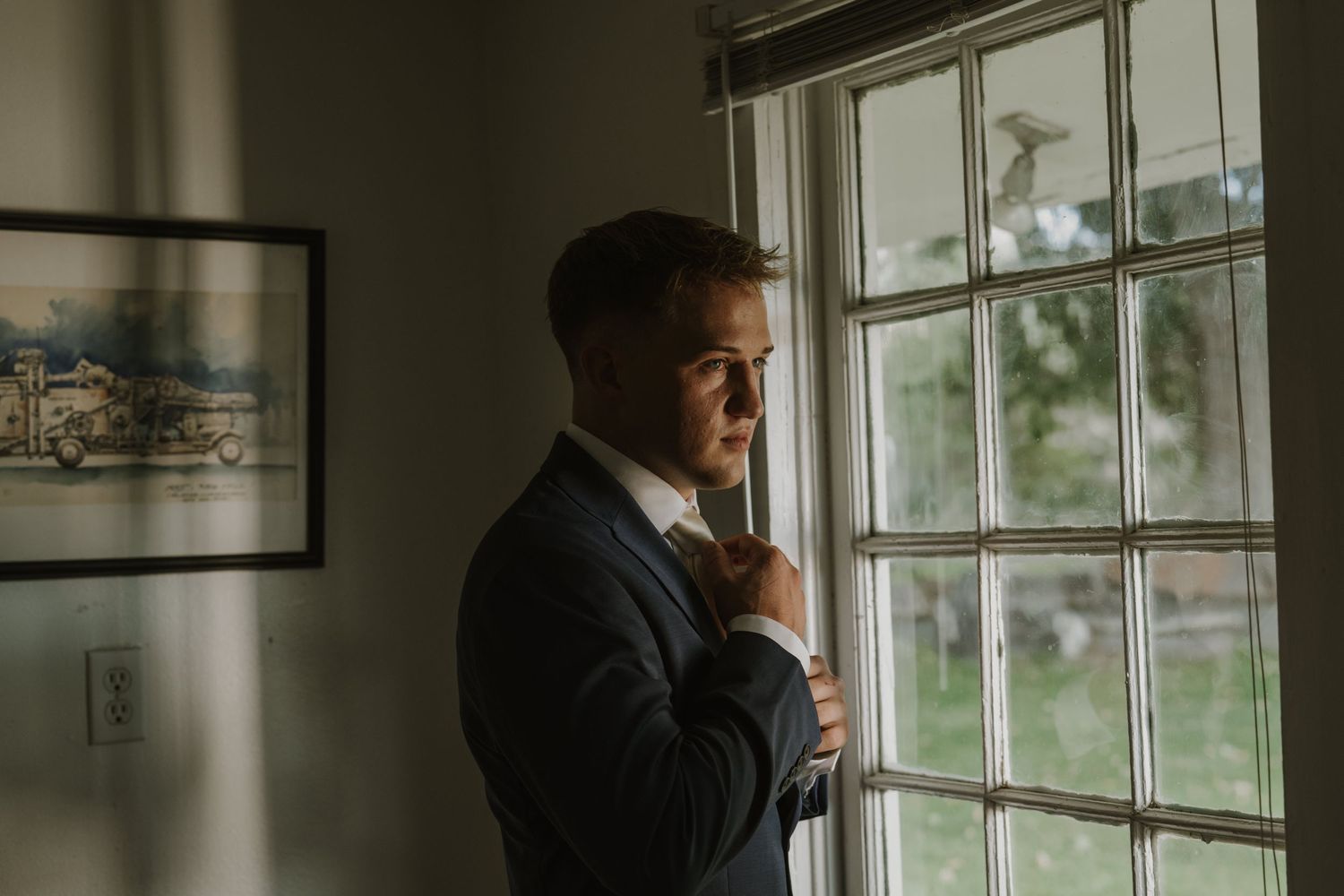 A figure in a dark suit looks pensively through a window in a bright interior room.