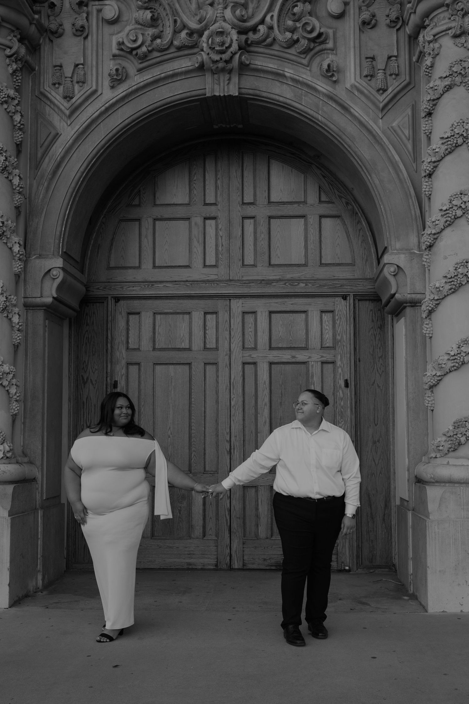A couple walks through ornate archways of a historic building in black and white.
