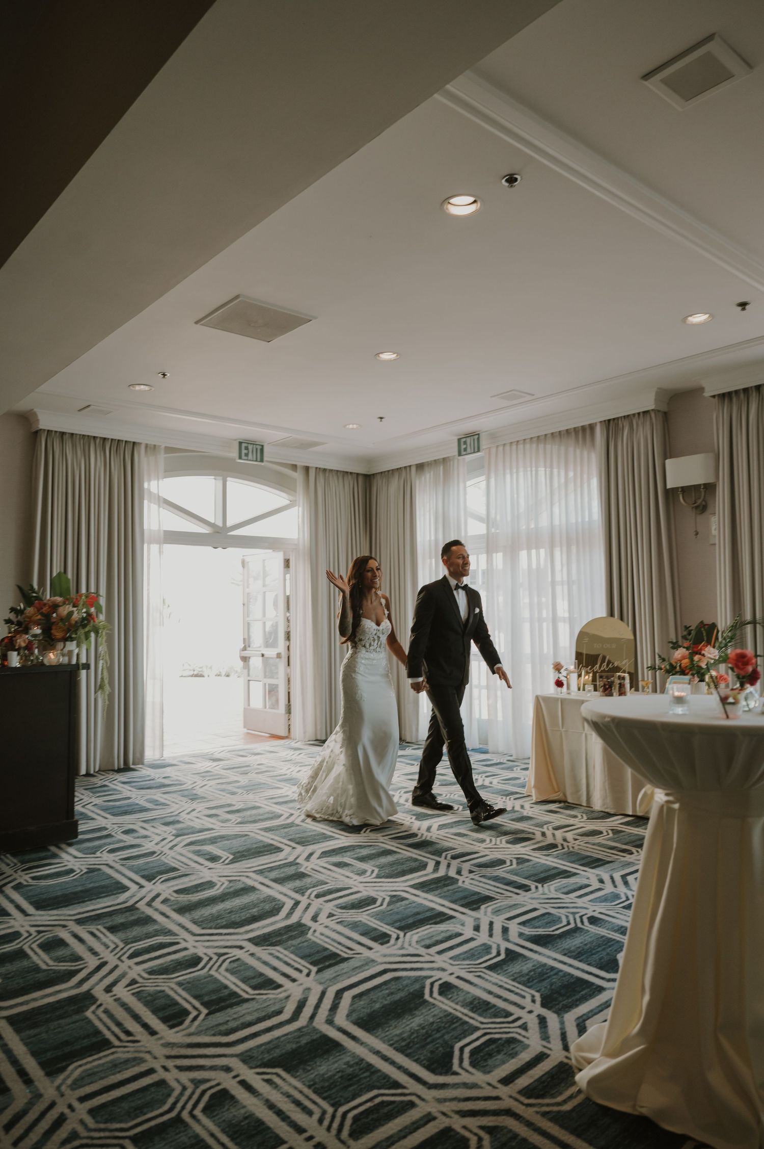 A couple makes their grand entrance into an elegantly decorated ballroom with geometric carpet patterns.