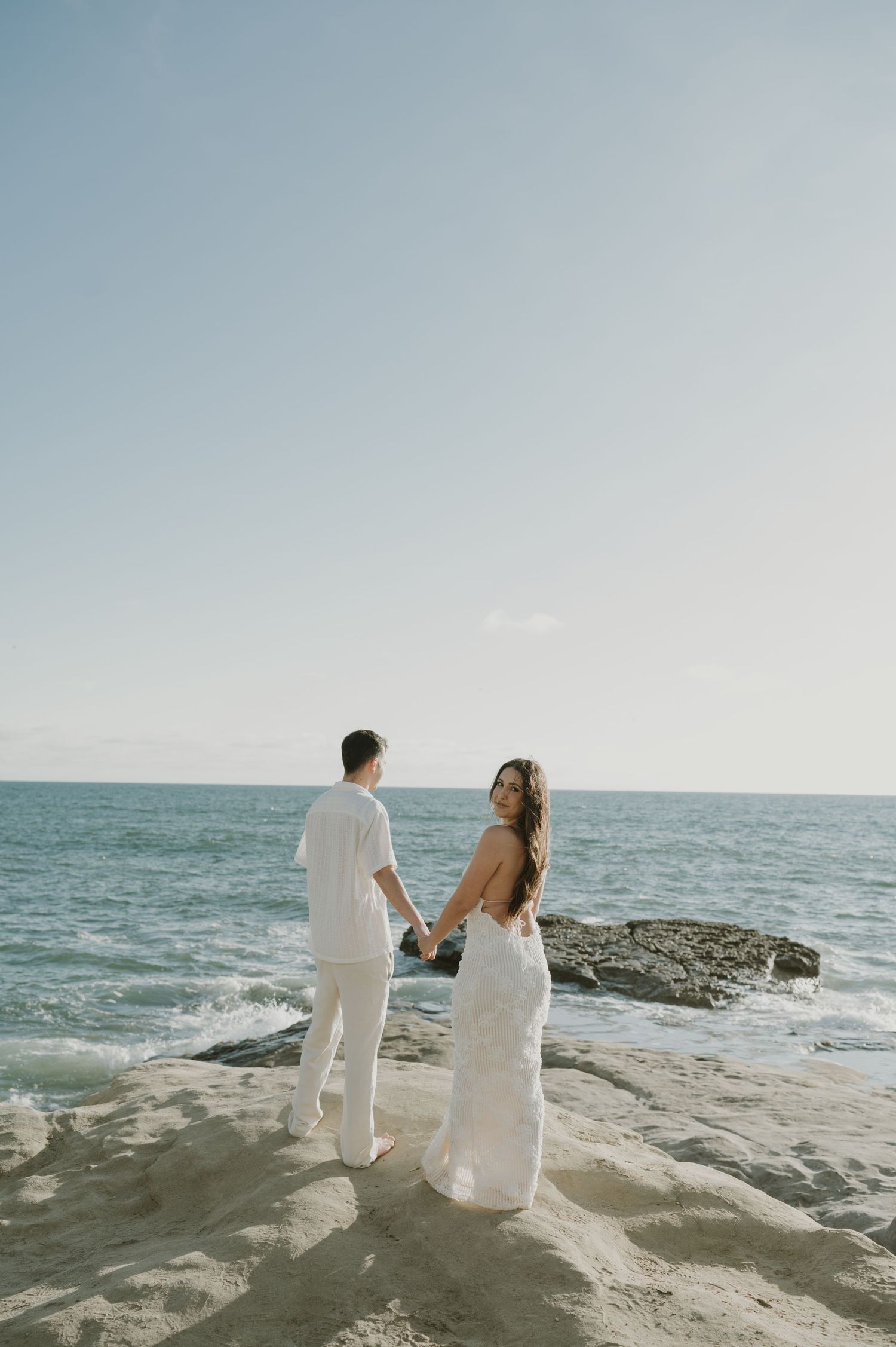 Couple in white attire sitting together on rocky coastal cliffs overlooking the ocean.