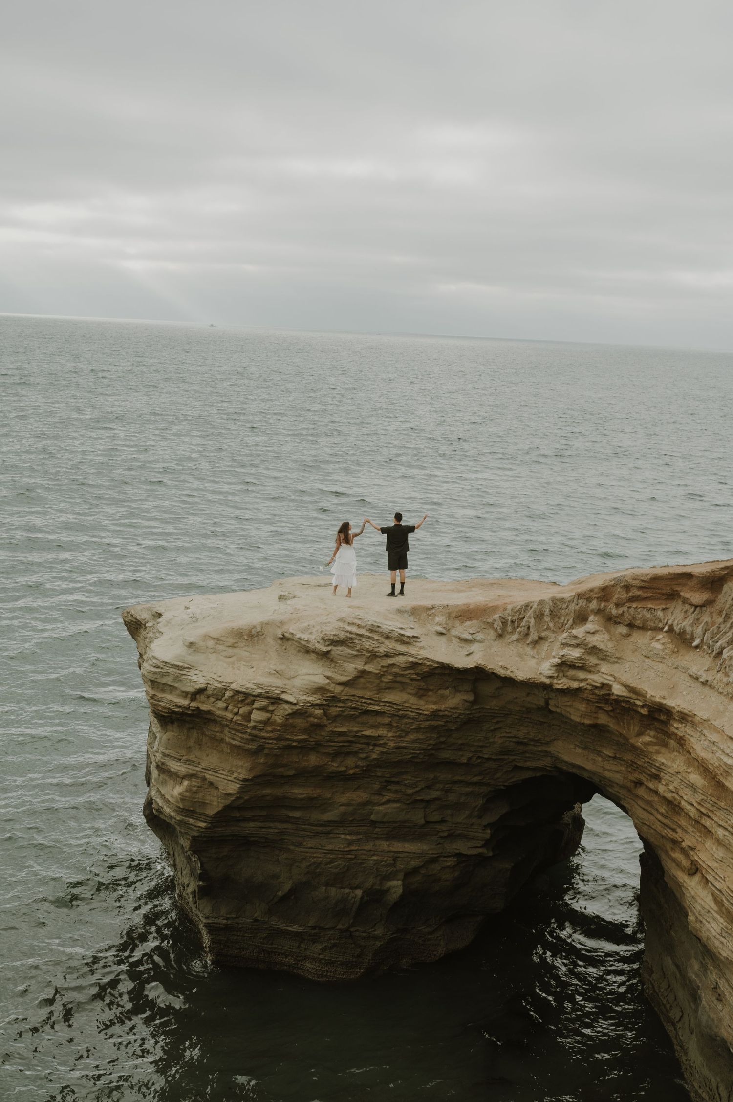 Two figures standing on dramatic ocean cliff overlook during overcast day.