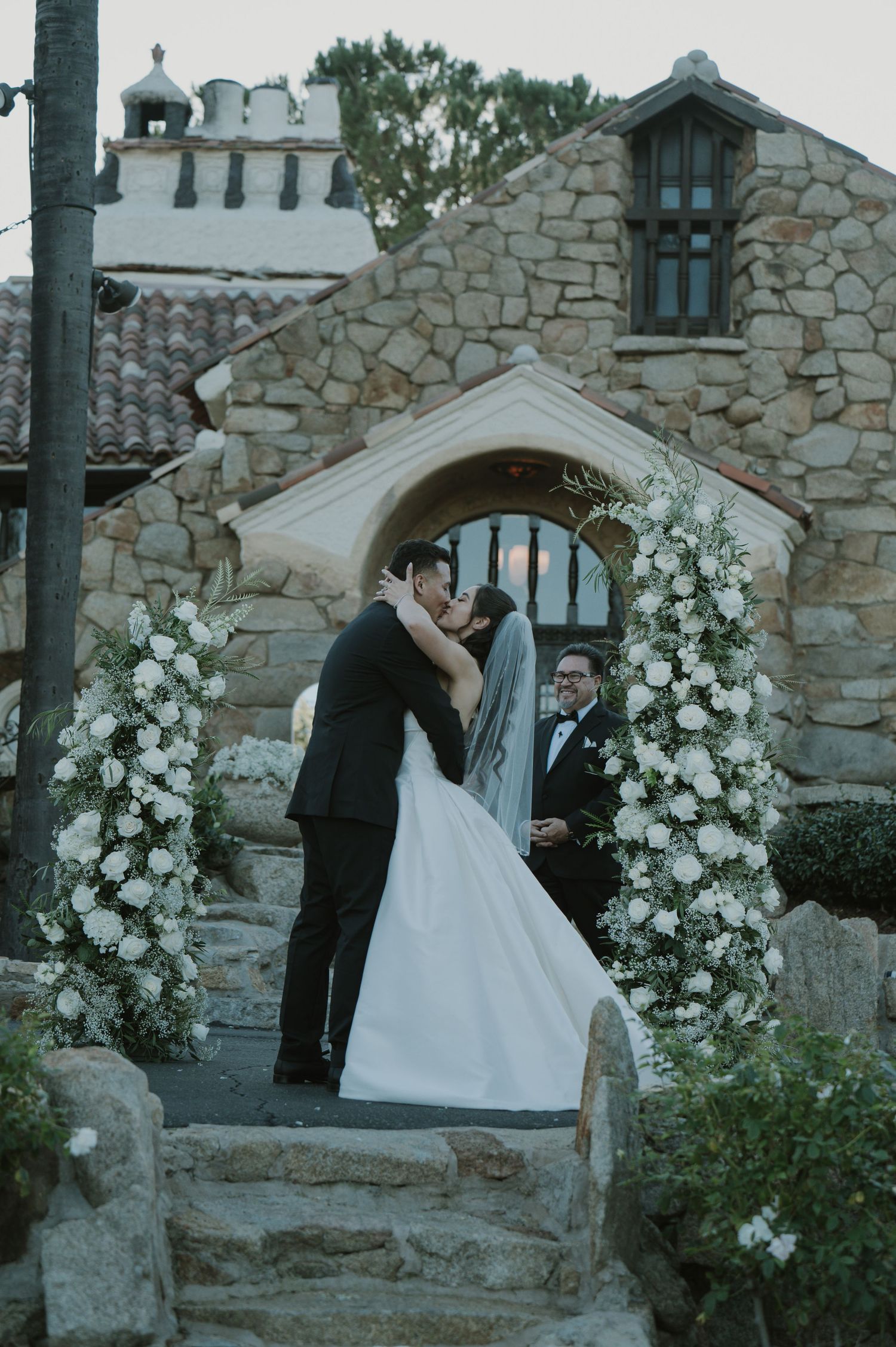 A romantic moment under a floral-adorned stone archway during a wedding ceremony.