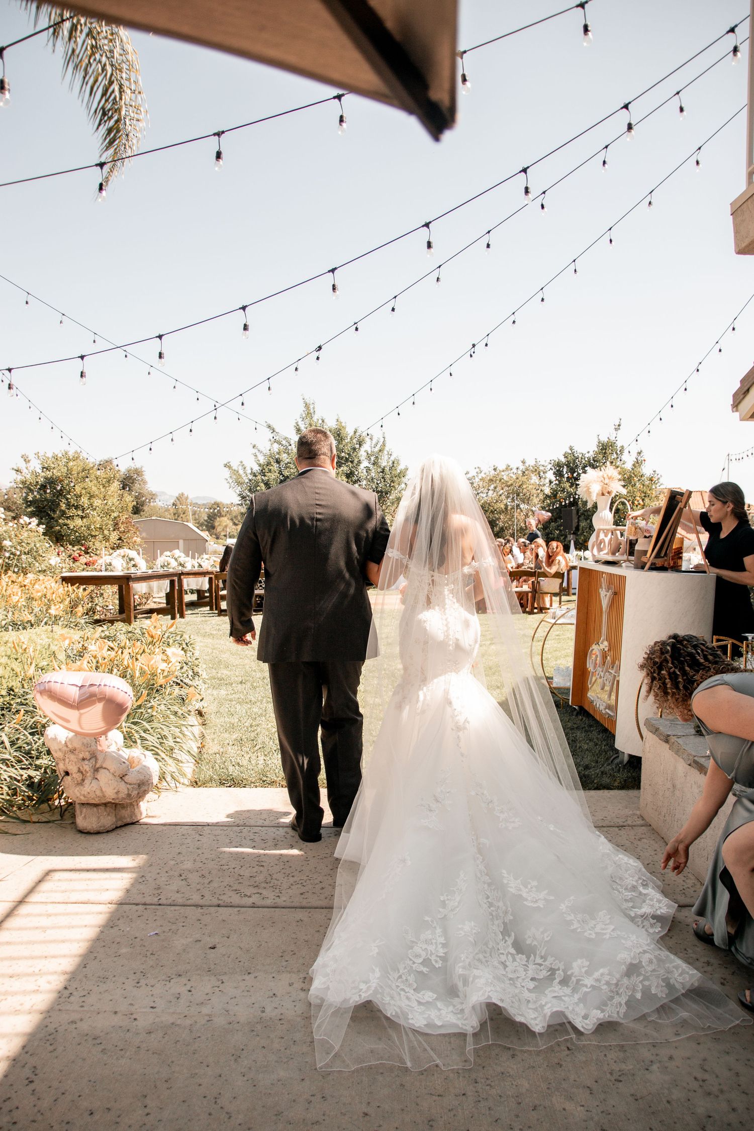 Bride and escort walking down outdoor aisle under string lights at sunset wedding ceremony.