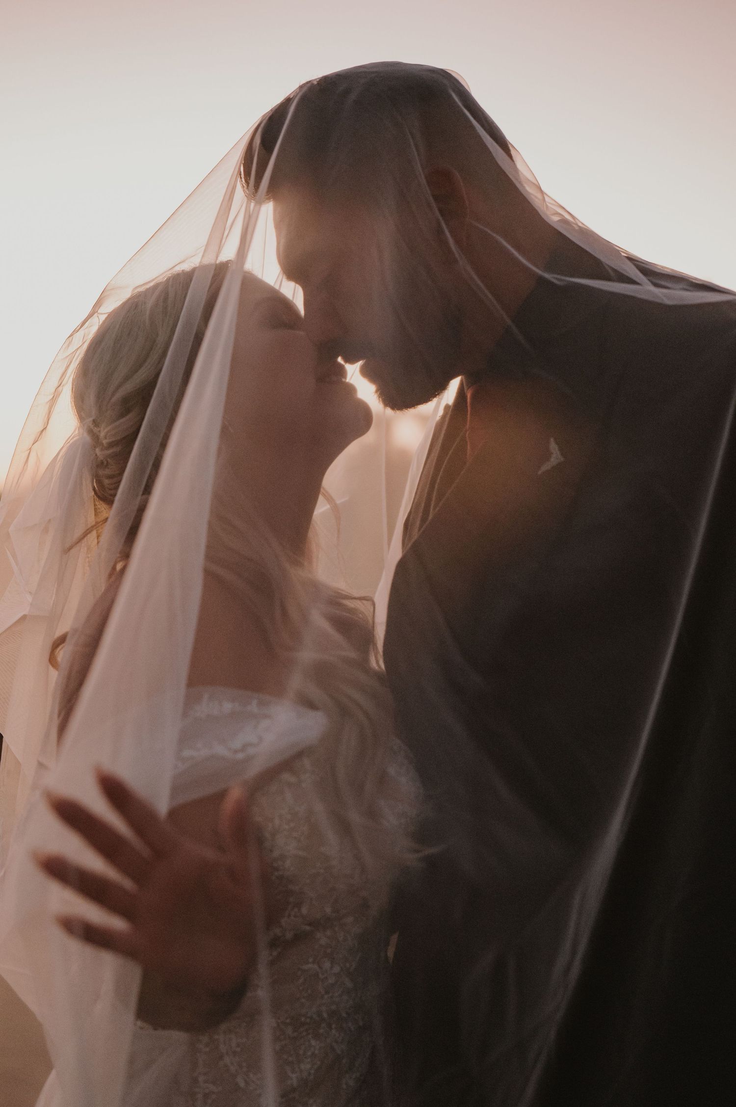 Romantic silhouette of bride and groom kissing under veil at sunset.