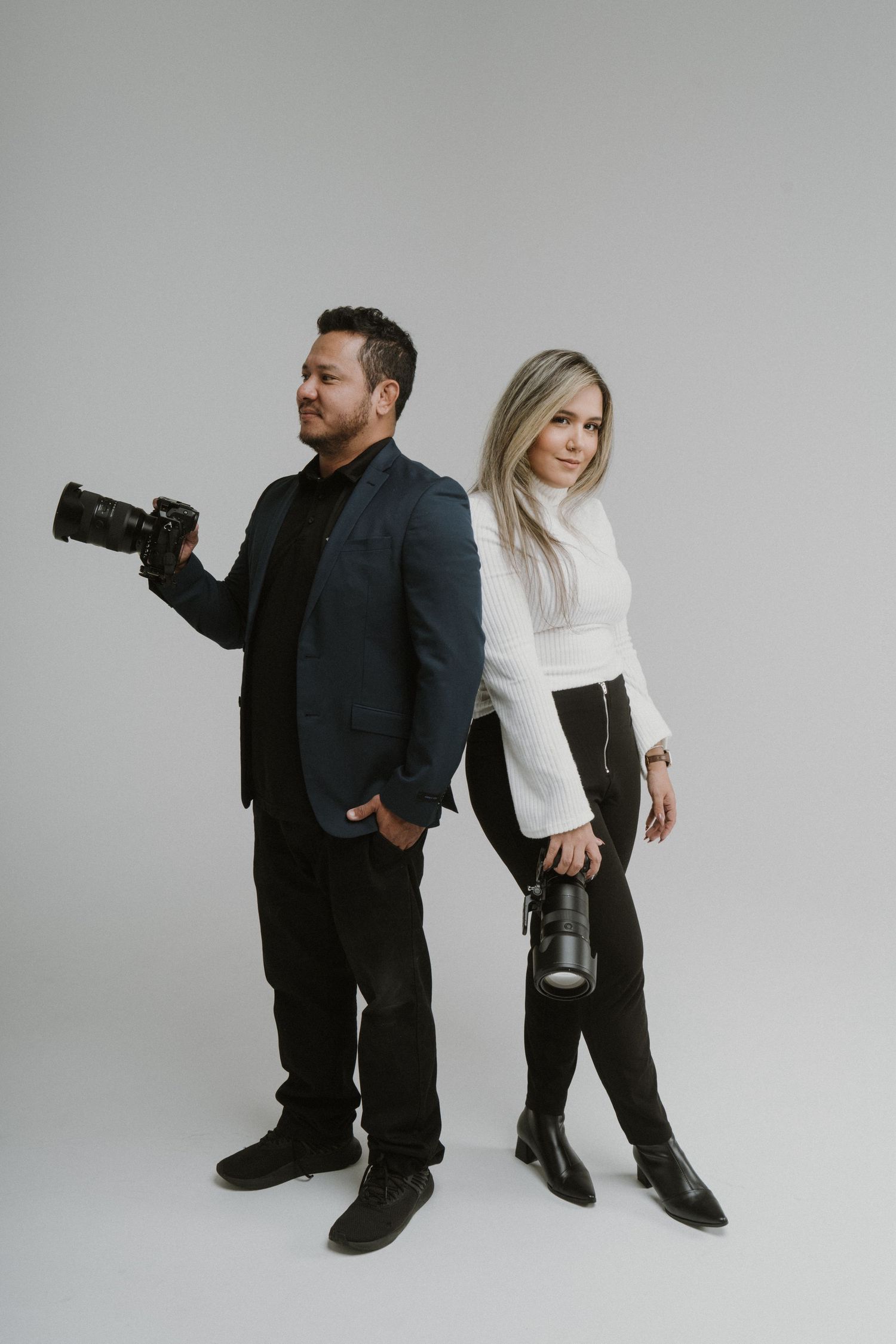Professional photography duo posing with cameras against grey backdrop.