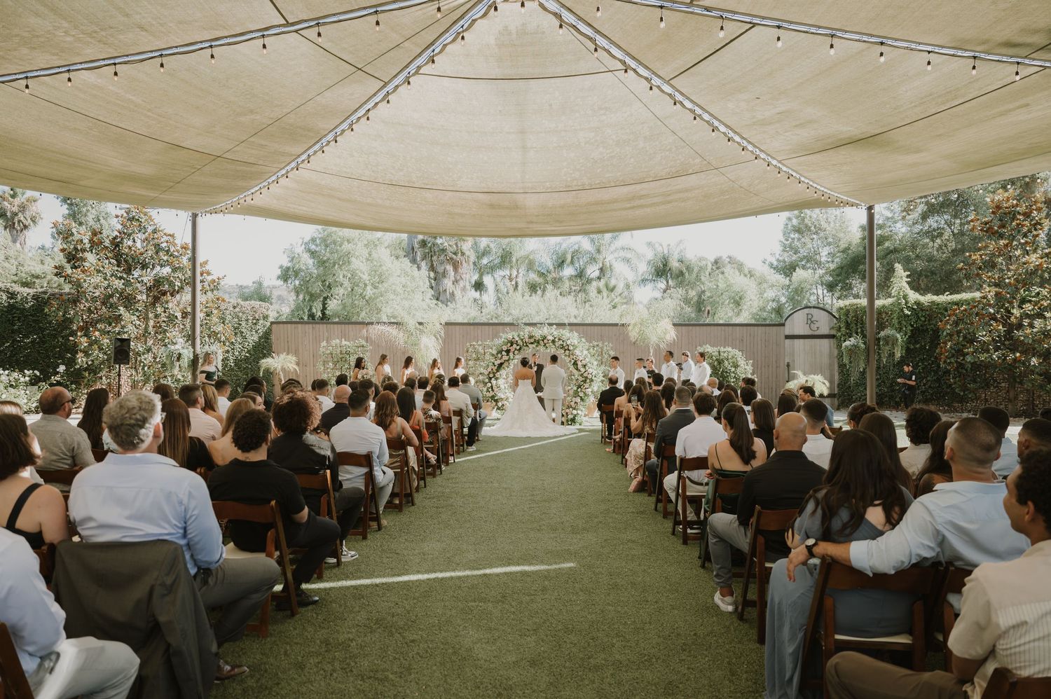 Wedding ceremony under a draped tent canopy with rows of guests seated on white chairs.