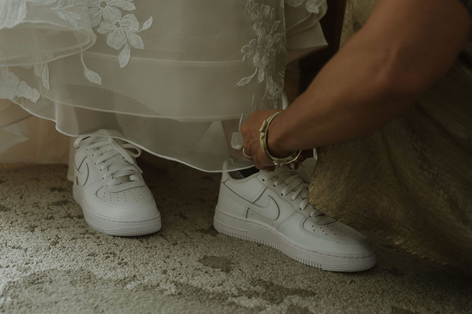 Bride wearing white Nike Air Force 1 sneakers under a floral lace wedding gown, tying shoelaces on concrete floor.
